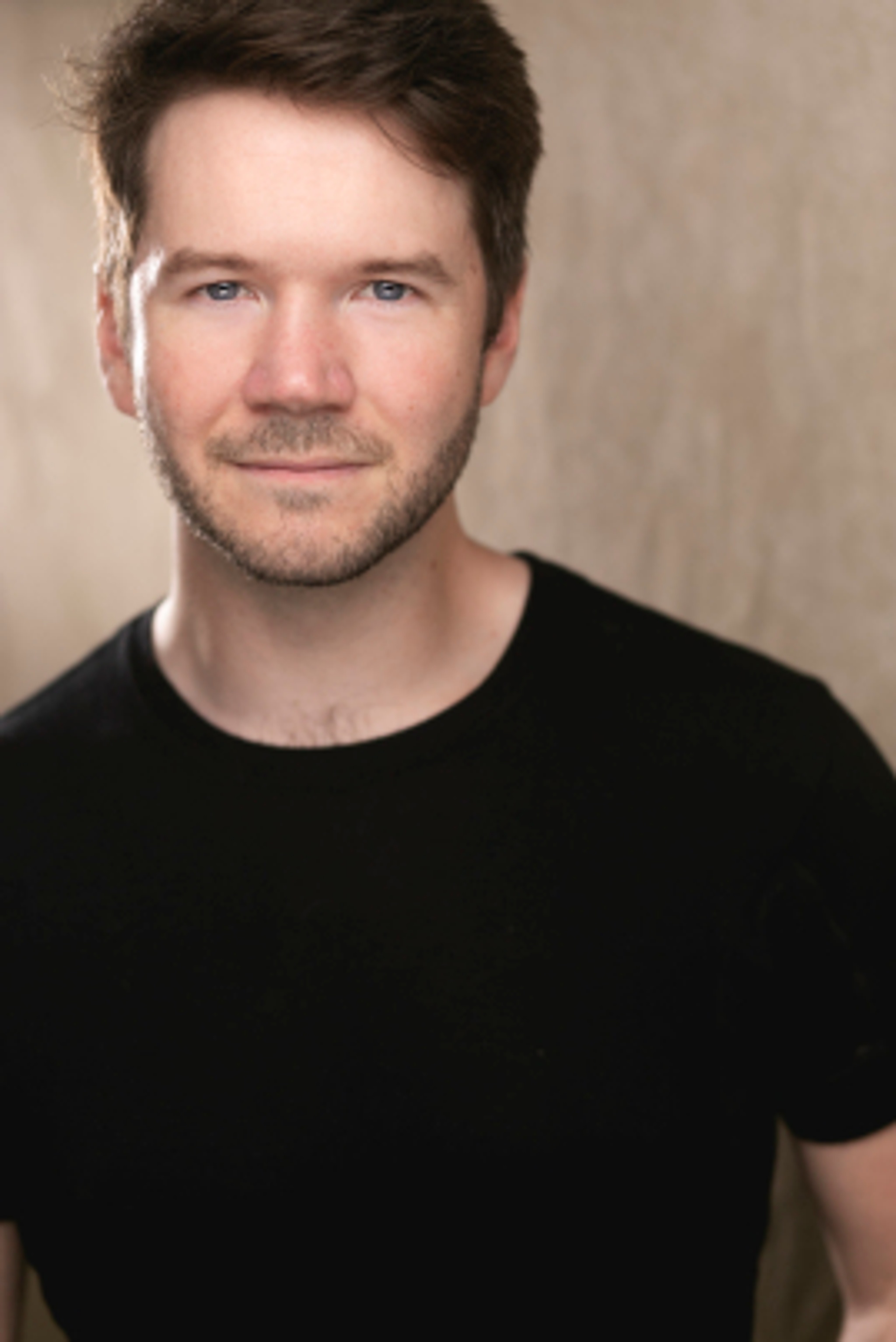 A portrait of artist James Caverly. Joey is a white man with brown hair parted to one side across his forehead. She smiles slightly and looks directly at us. He wears a black tshirt.