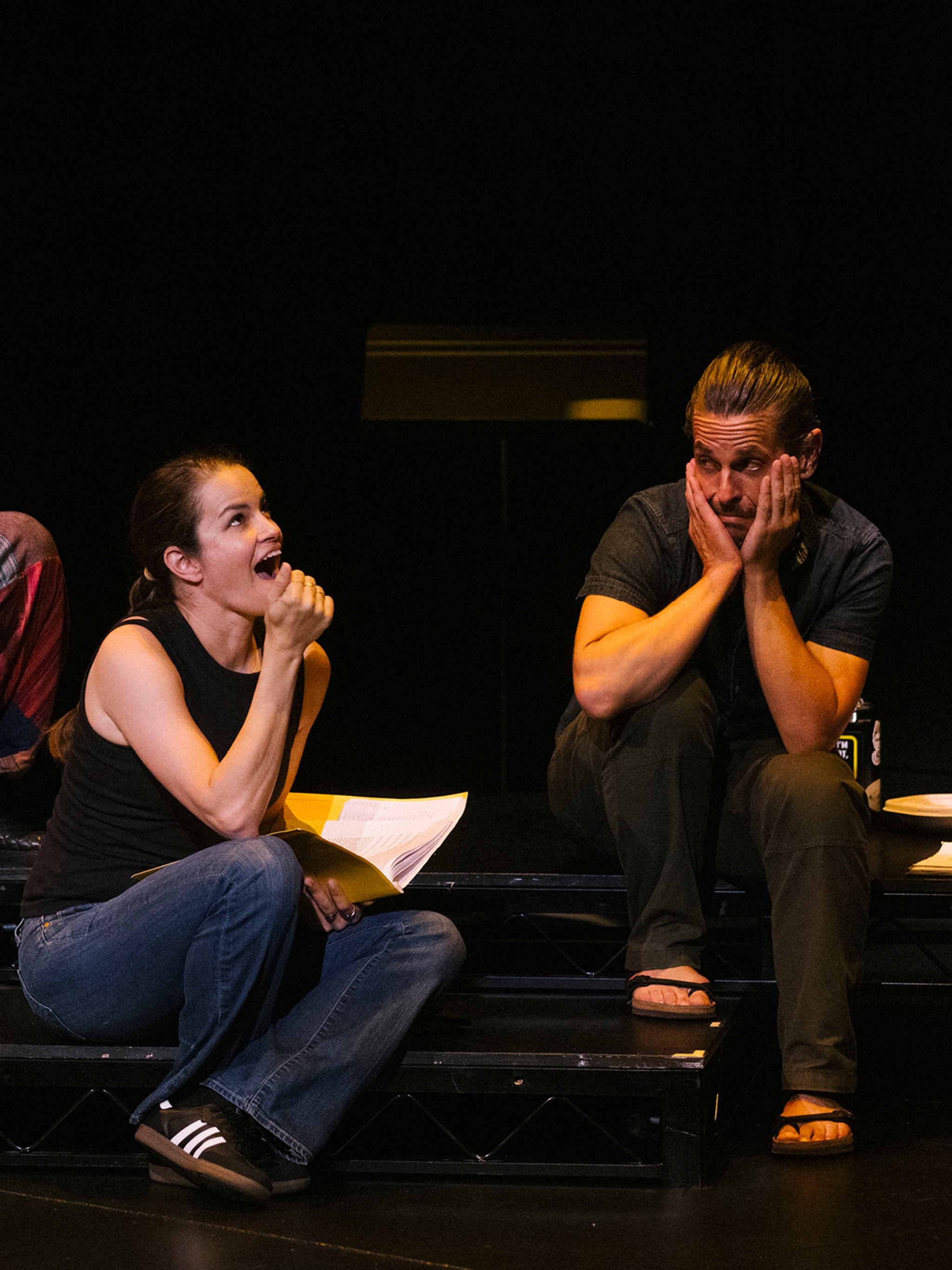 Two Deaf performers interact on stage in Garrett Zuercher's "Inside/Look." Both sit on the ground, the woman looking up with her mouth open in amazement while the man looks at her skeptically from the side, with his hands held to the sides of his face. 