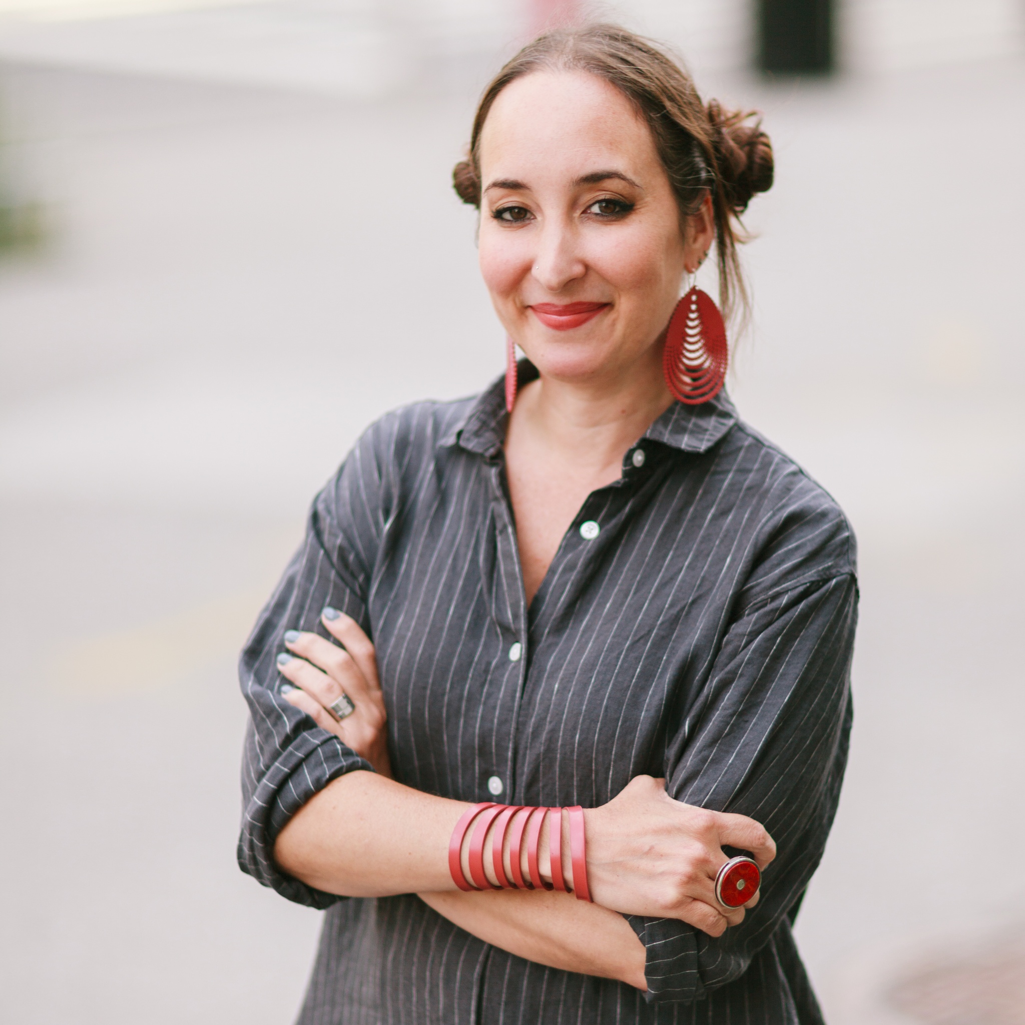 A photo of Marlène Ramírez-Cancio with arms crossed over her torso and wearing a gray shirt with pinstripes. She wears red jewelry including a bracelet with multiple straps up her lower forearm. 