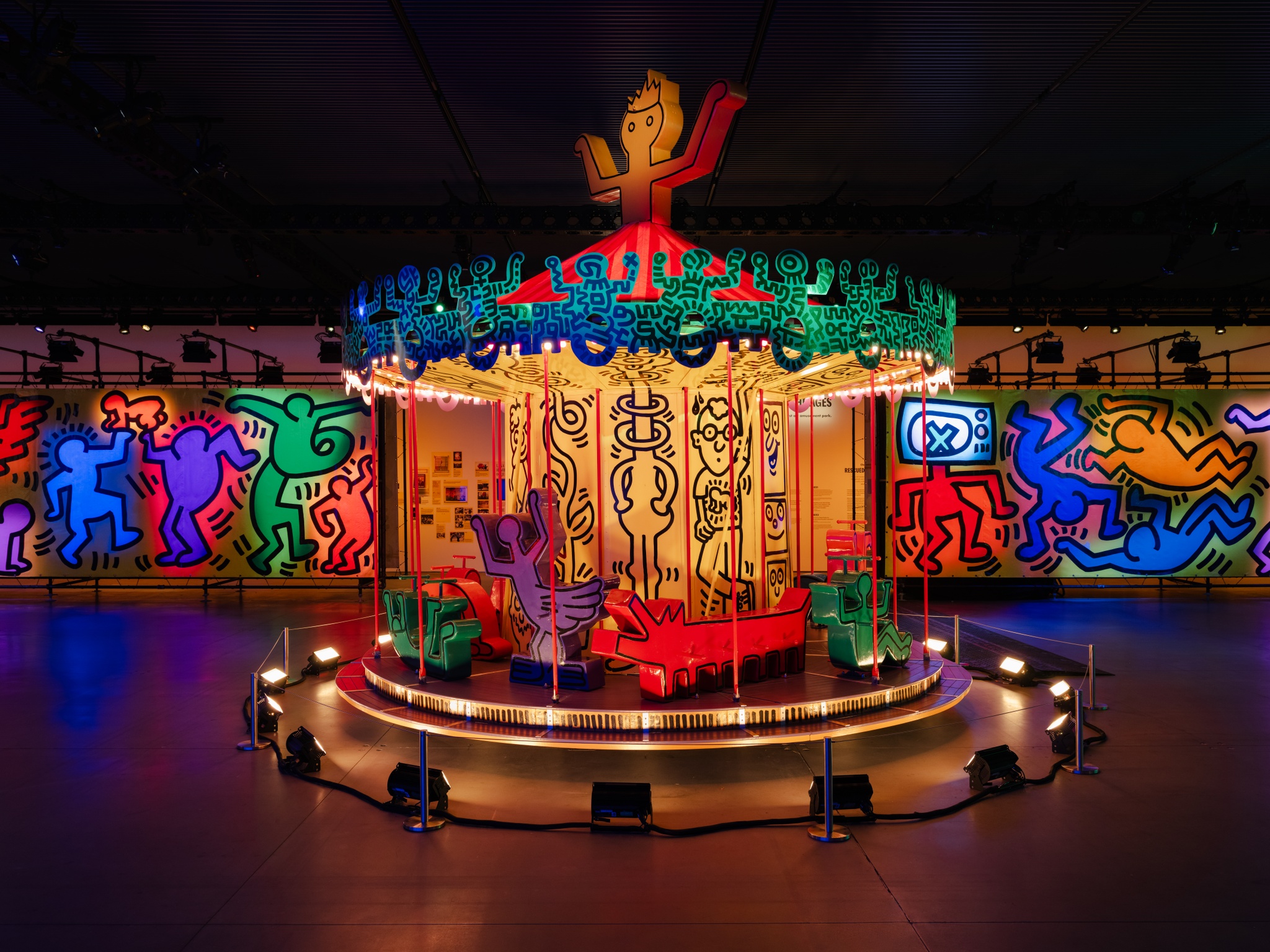 An art carousel painted bright yellow and green by Keith Haring, with his characteristic outlined shapes of jubilant humans decorating the ride.