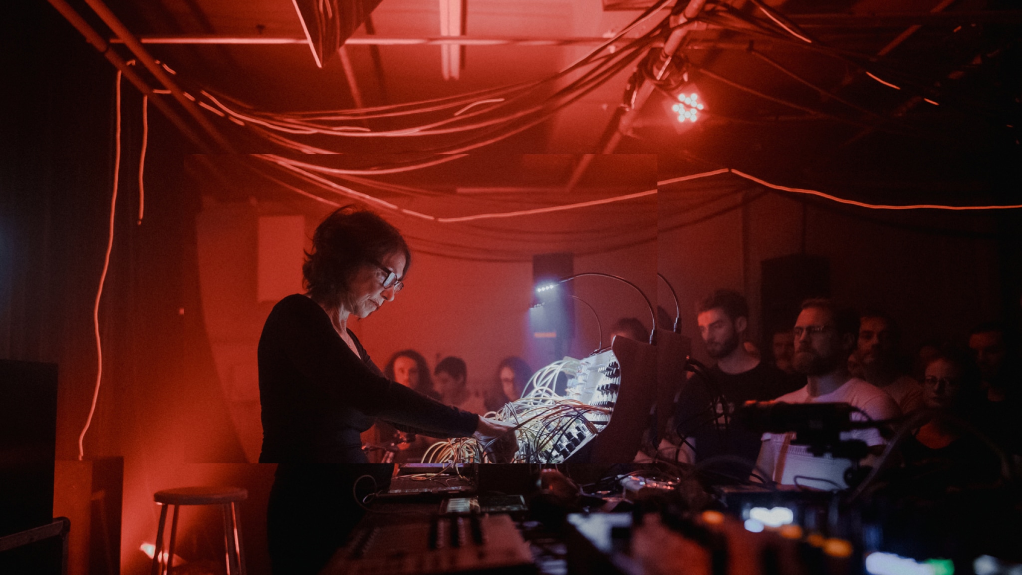 Musician Suzanne Ciani, a white woman with short wavy brown hair, is seen in profile manipulating a sound board. The small space is lit by red light and an audience is gathered on the other side of the sound board. 