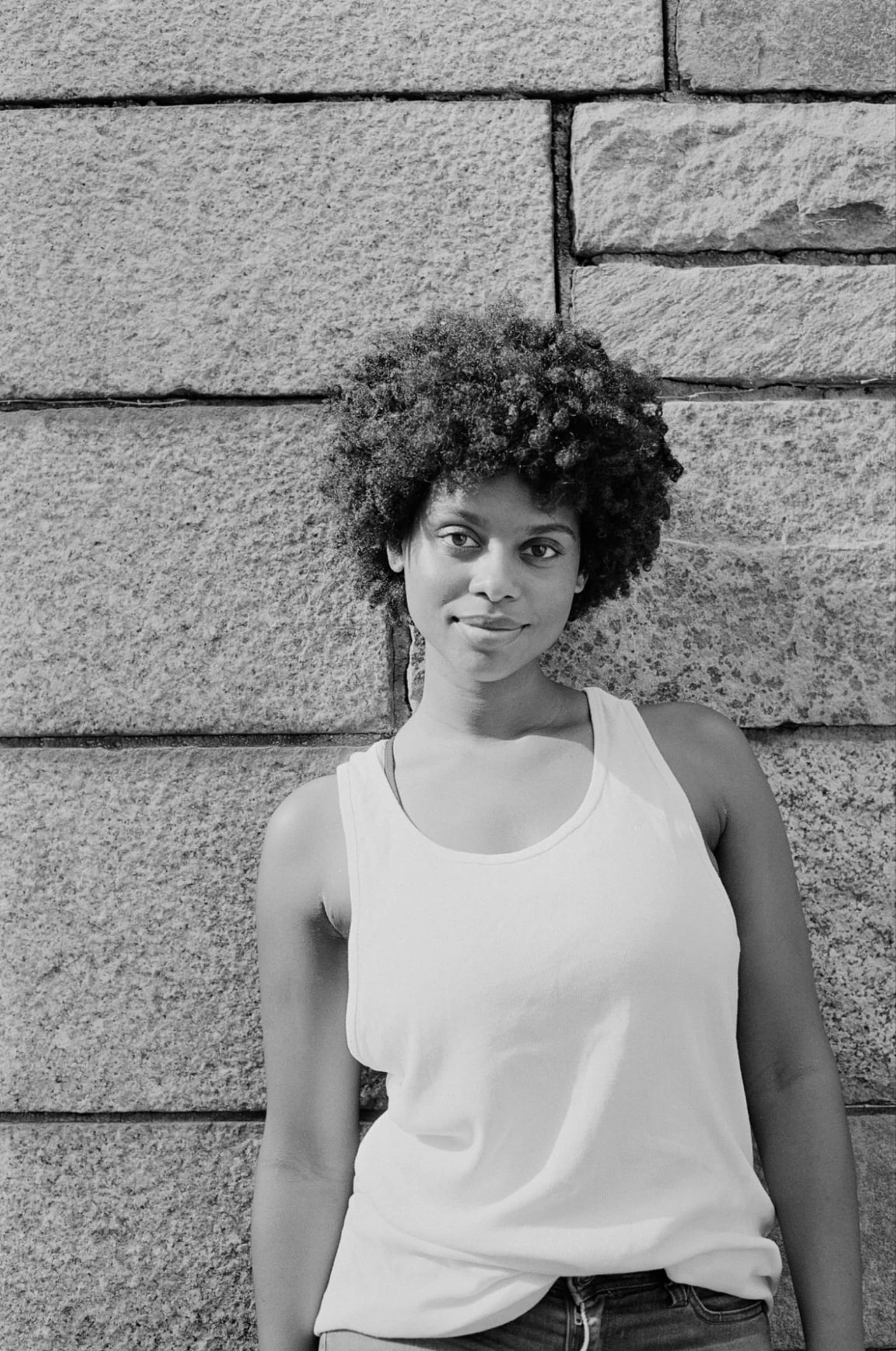 A black and white portrait of performer Kelah Winfield, a Black woman who wears a white tank top and slouches slightly against a stone wall. She looks directly at us and wears her hair in a short Afro. 