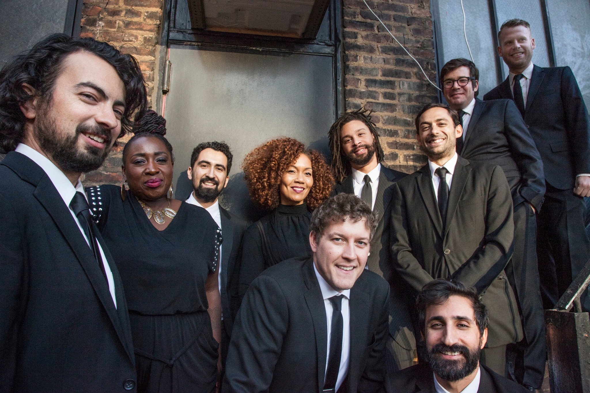 Ten members of the band The Cool Rulers dressed in suits and ties and black dresses, lined up along a staircase beside a brick building façade