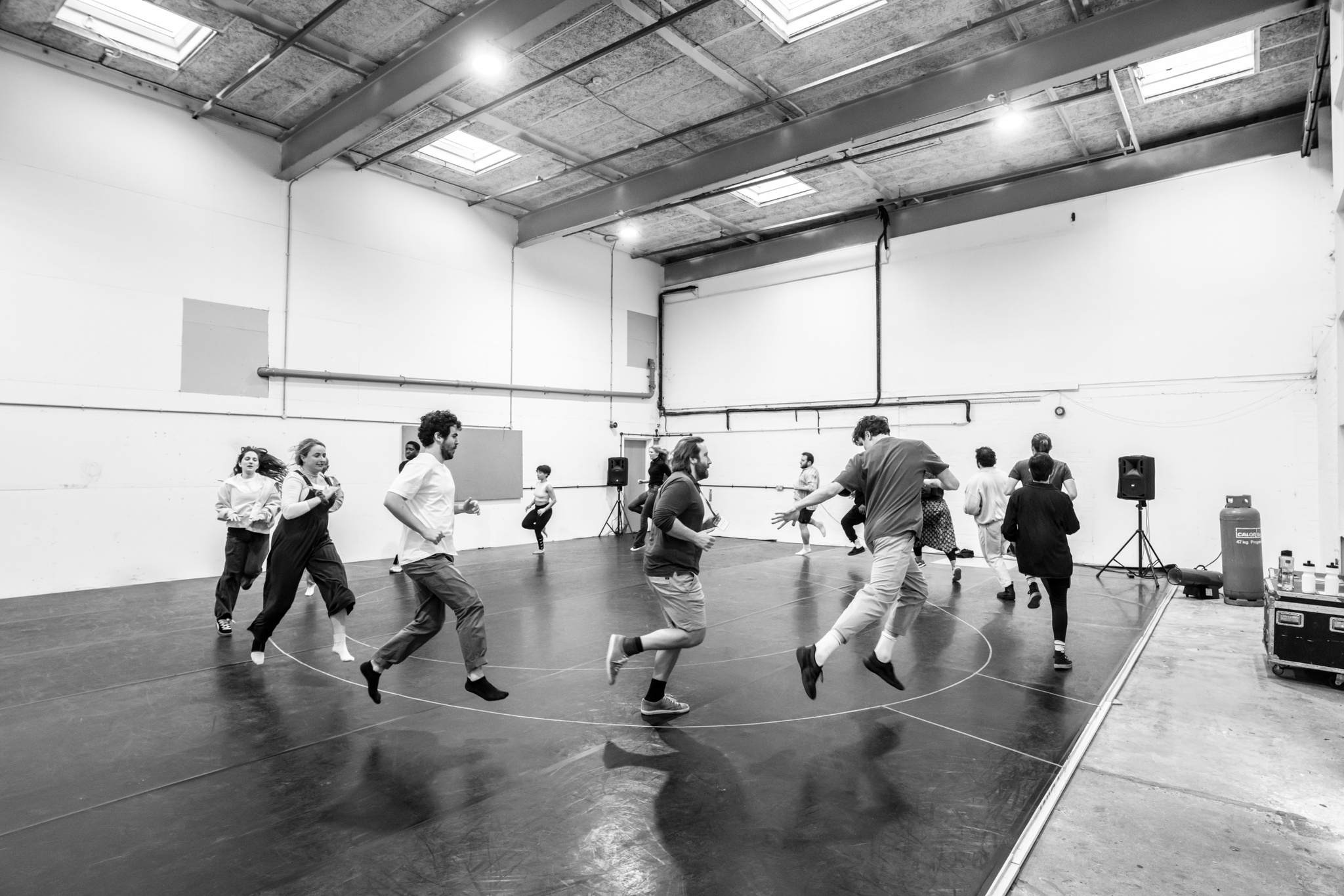 15 King Lear cast members run in a circle in rehearsal. They leap and look behind them, with smiles on their faces. 