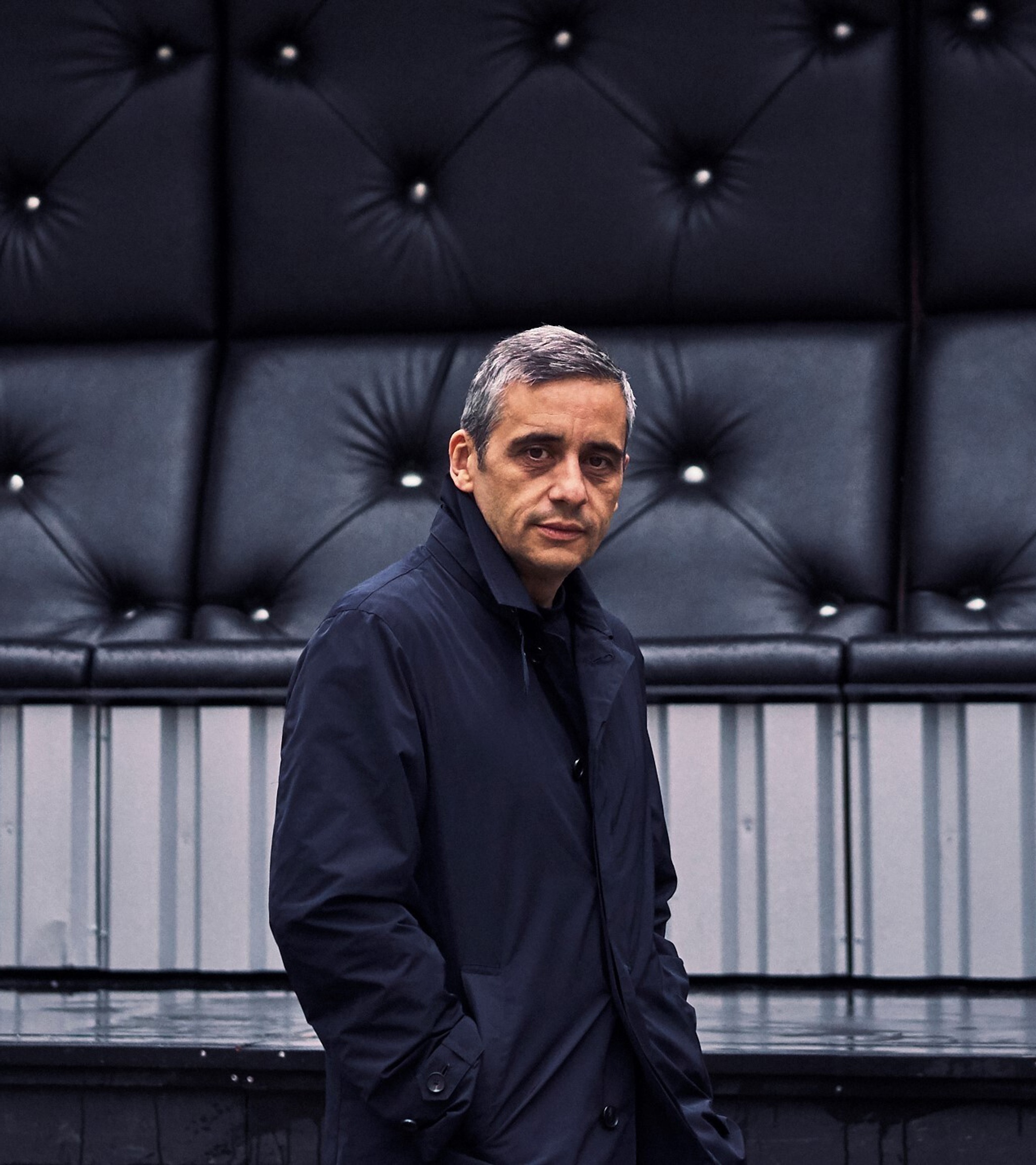 A portrait of Alex Poots, The Shed's artistic director. Alex, a white man with salt and pepper hair, stands against a bank of outdoor seating. He is wearing a navy blue coat buttoned to his neck and stands with hands in his pockets, turning slightly to look directly at us. 