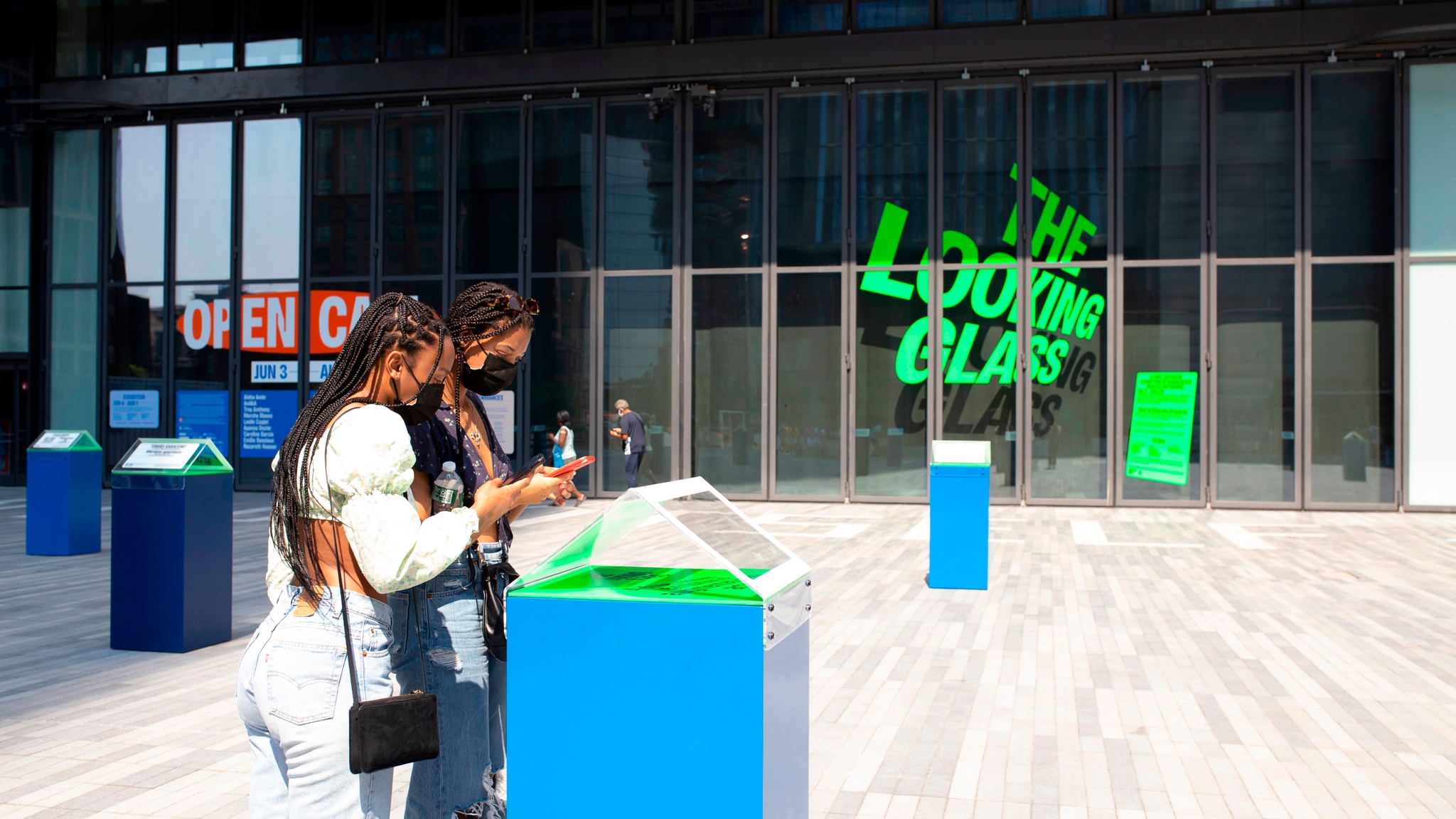 Two people with long braids lean over a bright blue plinth used to activate the exhibition The Looking Glass on a sunny outdoor plaza