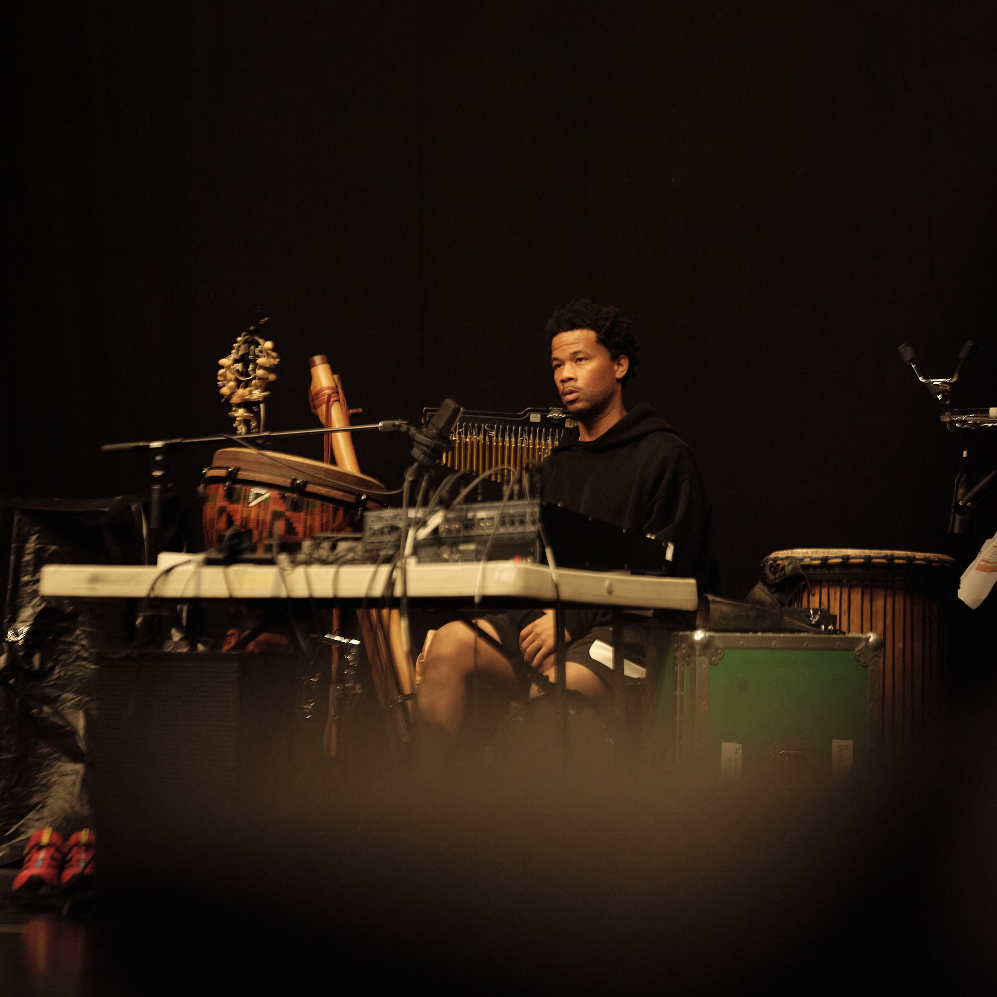 Musician Munir Zakee, a Black man, sits amongst a set up of various instruments in a black-box rehearsal theater. He wears a black sweatshirt and has his gaze trained on rehearsals happening out of frame of the image.