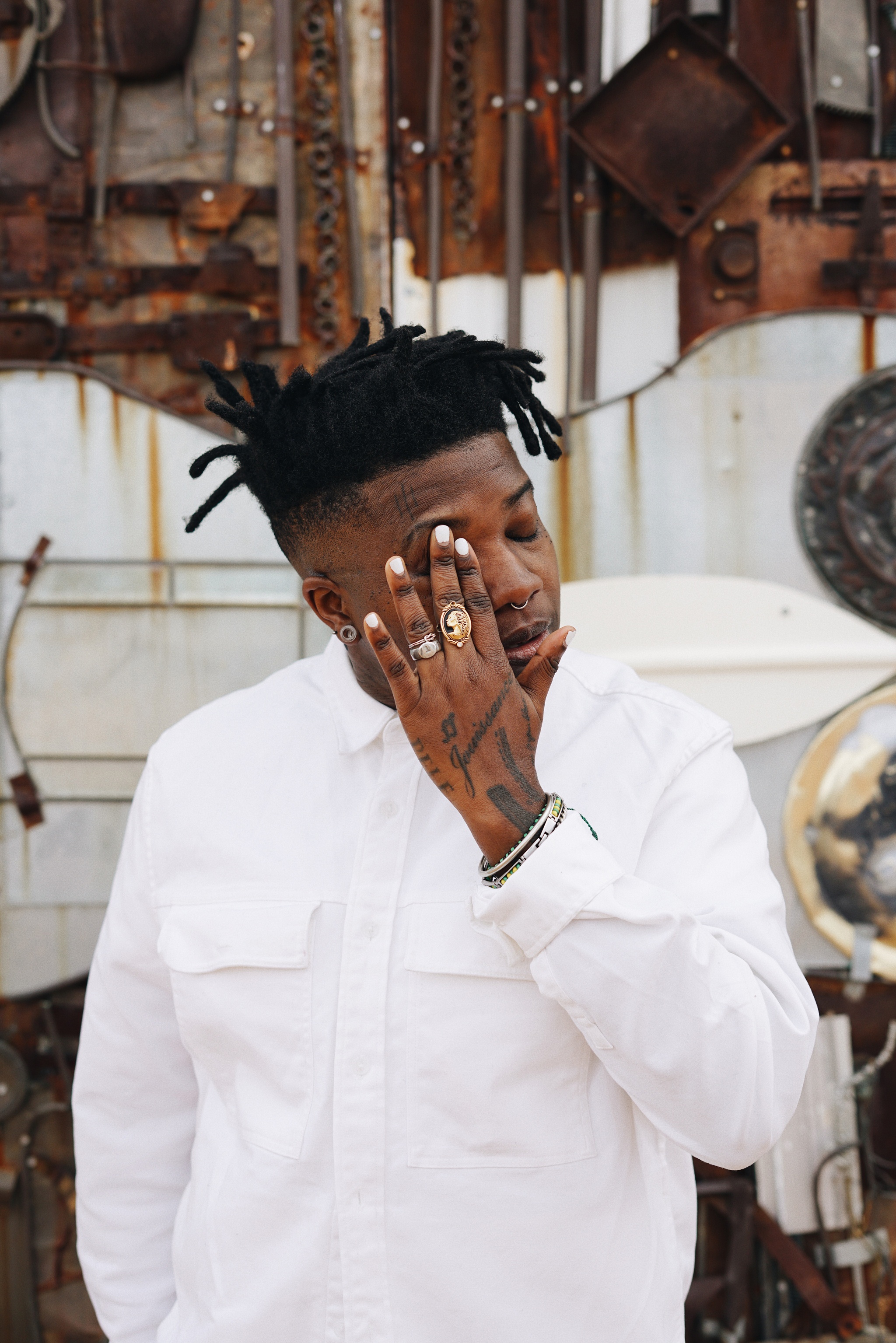 A photo of artist Tiona Nekkia McClodden against a background of rusted metal elements. McClodden's face is obscured by her left hand held up to her right eye and she wears a white shirt. 