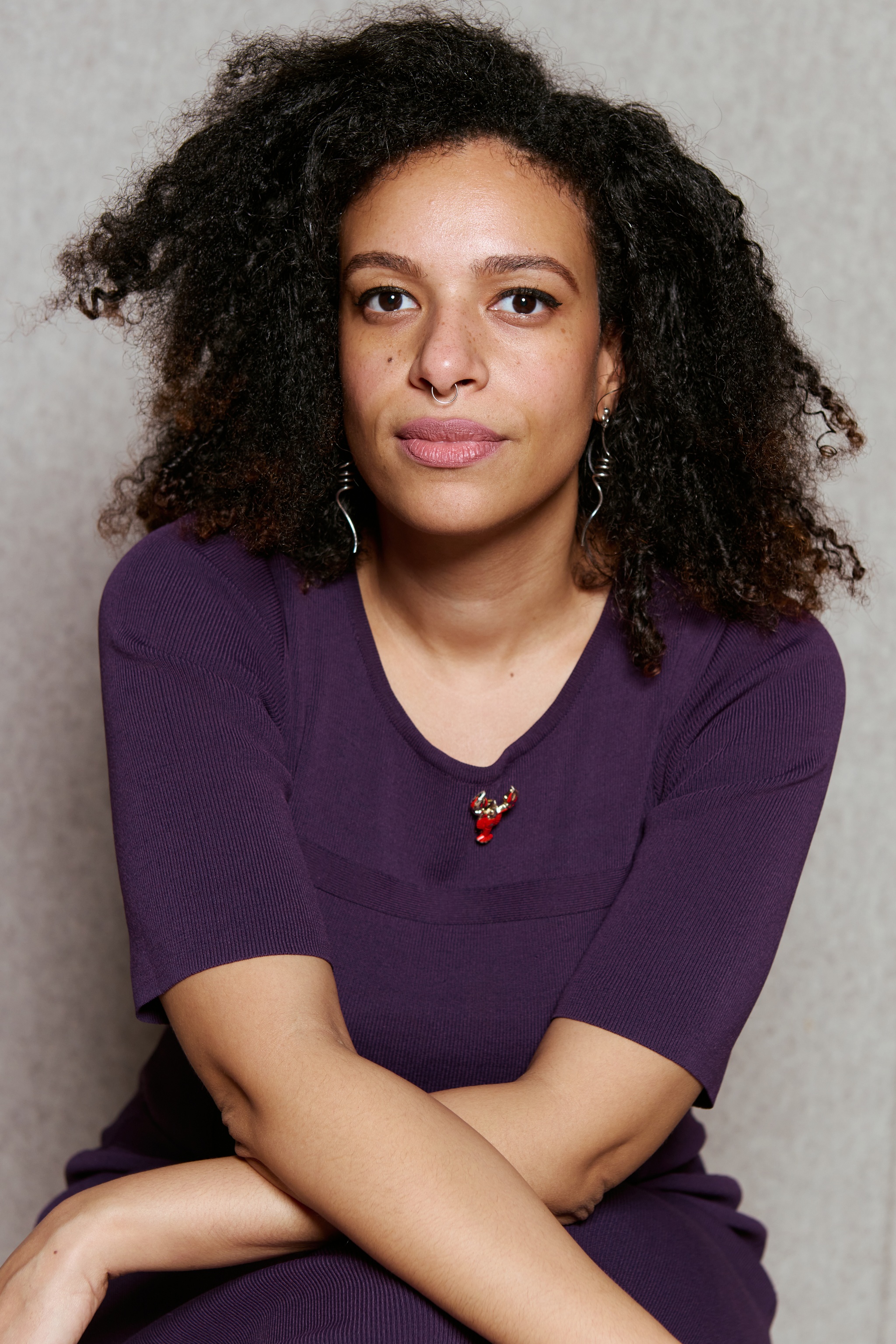 A portrait of artist Yelaine Rodriguez. She has curly, long brown hair and wears a purple dress. She poses against a beige felt background with her arms crossed over her knees, leaning forward slightly, and looking intently at us. 