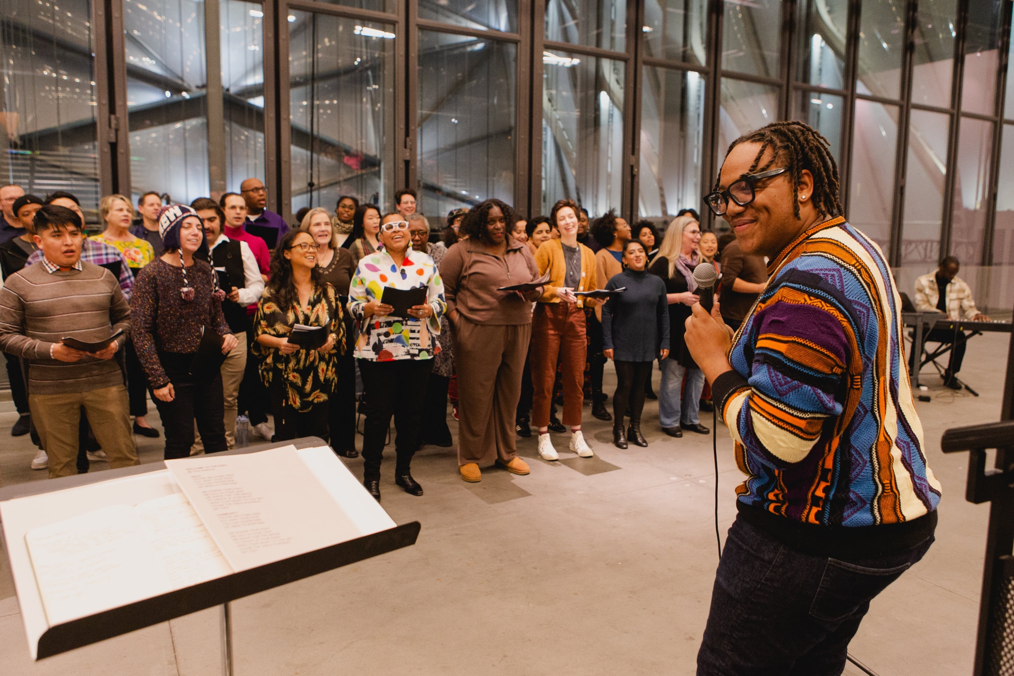 The Fire Ensemble choir rehearses in an event space at The Shed. To the left, the choir's artistic director Troy Anthony stands holding a microphone facing the choir, his head turned to the side with a broad smile on his face.