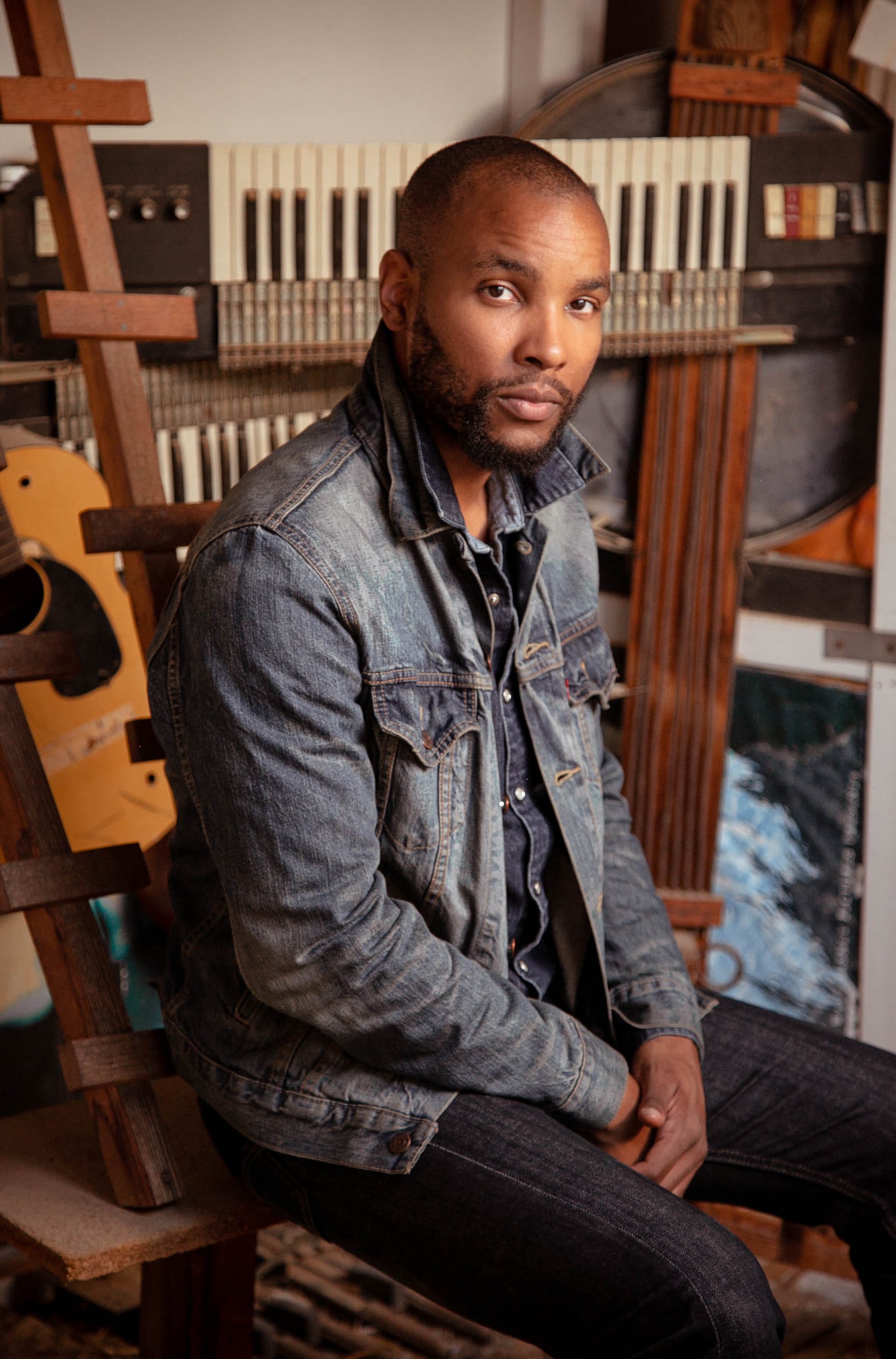 A portrait of actor Alani iLongwe in a studio space. Alani, a Black man with a shaved head and light beard and mustache, turns to look at us slightly from the side. He wears a jean jacket and sits in a relaxed pose with hands in lap. Behind him are instruments that seem to be in storage: a keyboard and a guitar. 