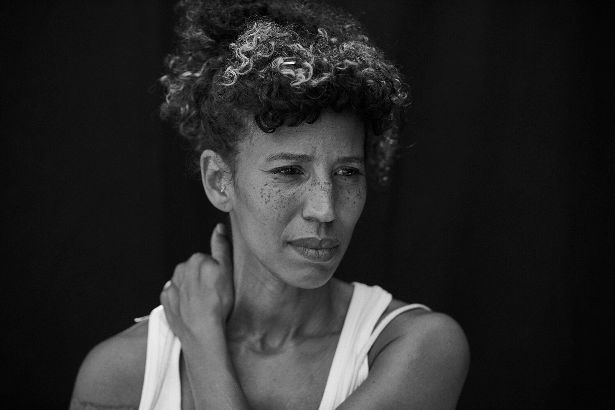 A woman with curly hair pulled up and back behind her head. She holds her neck with her left hand and has a look of concentration on her face. 