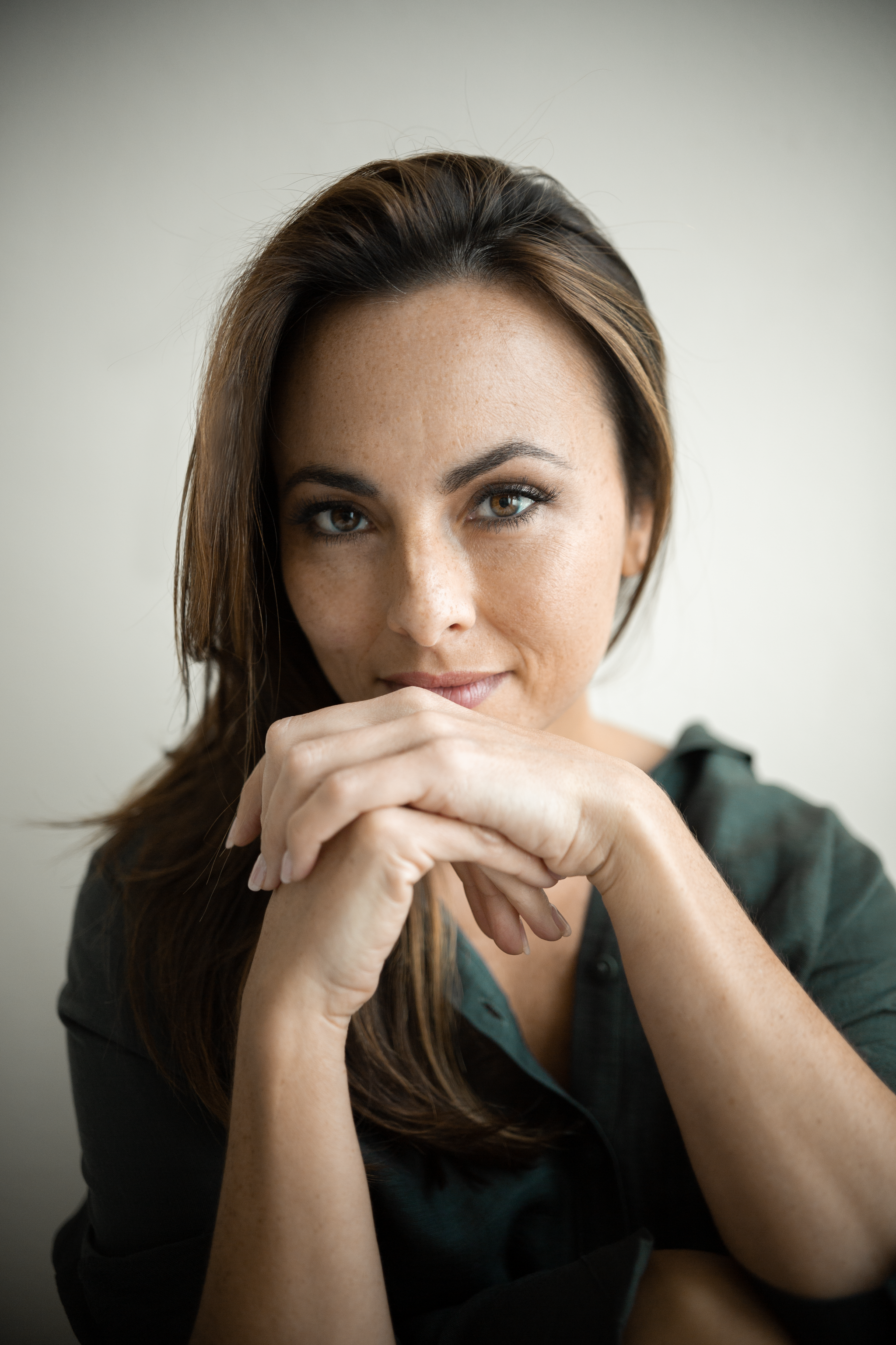 The mezzo-soprano Isabel Leonard, a white woman with straight brown hair pulled to one side of her head, looks directly at us, resting her face on her clasped hands. She wears a dark green blouse.