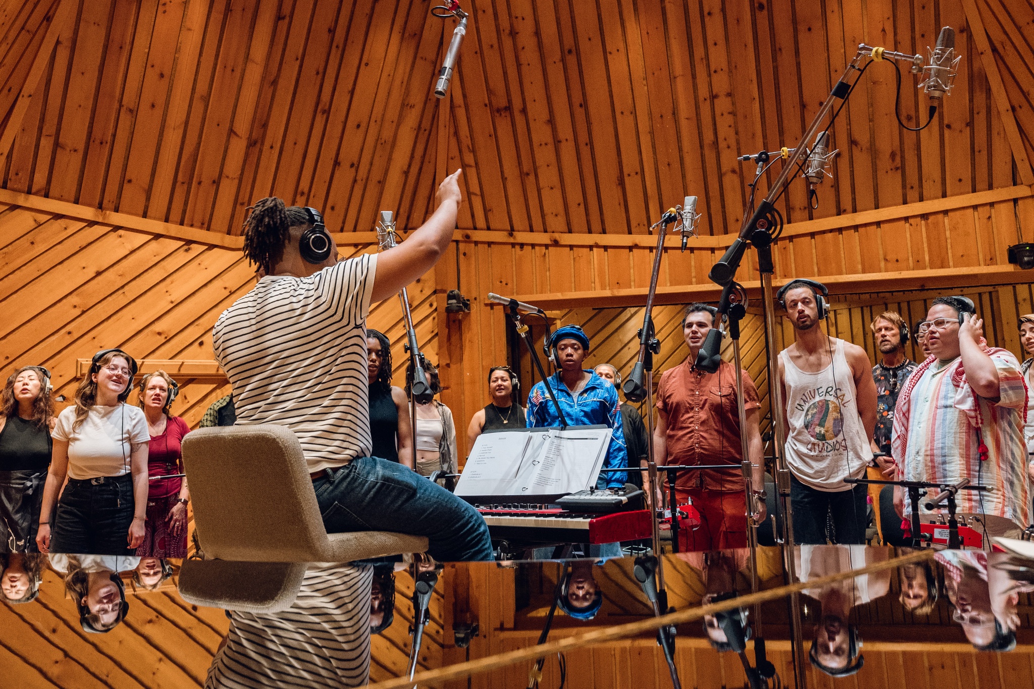 Troy Anthony is seen from behind, his arm in the air leading a choir as they sing in a vaulted, wood-lined recording studio. The choir stands in front of Troy, facing him and us. In the foreground, Troy is reflected in the smooth black surface of a piano.