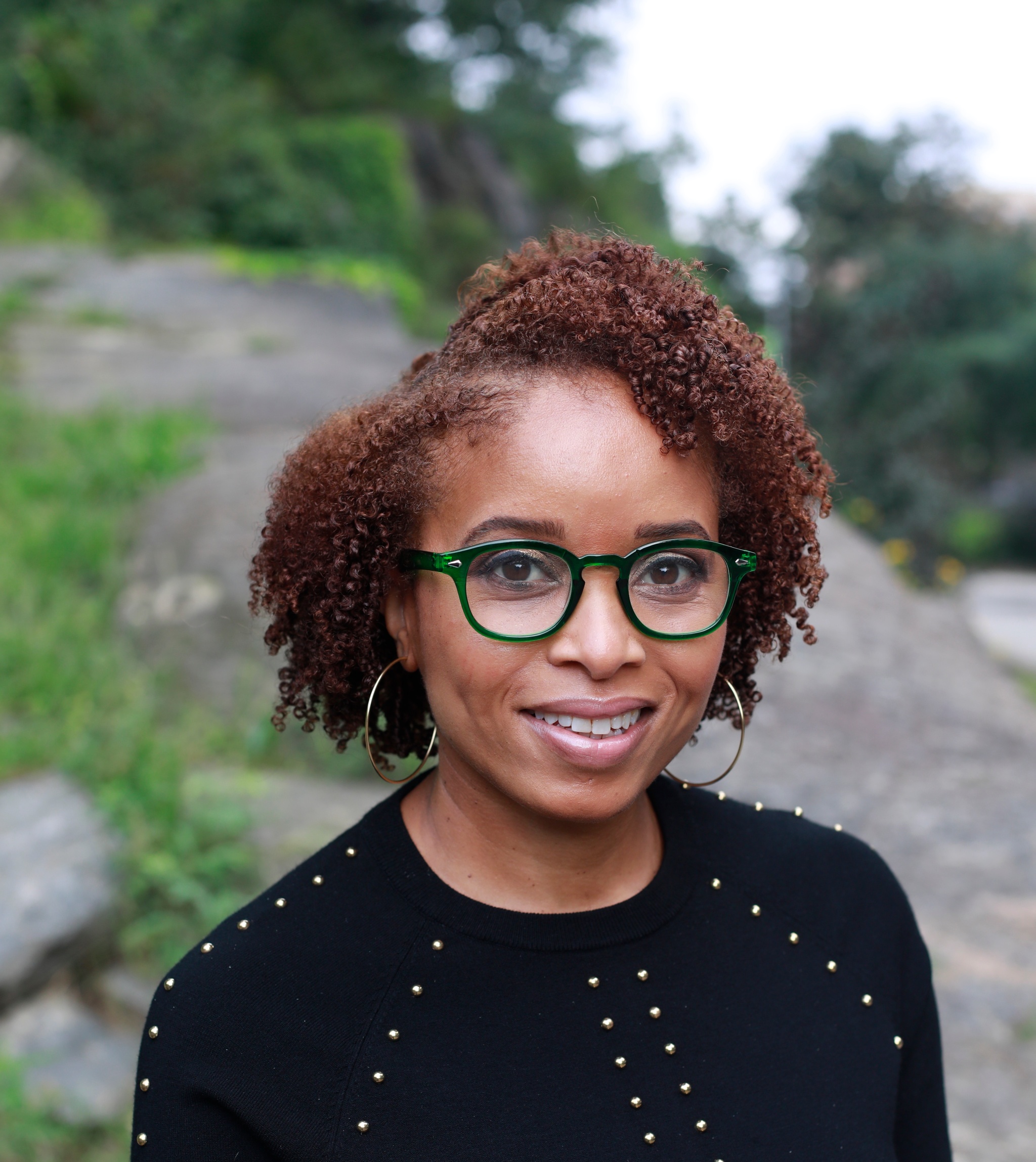 A photo of Nicole Fleetwood, who has short hair parted on the right side of her head and wears glasses with emerald green frames. She poses outside with a rocky outcrop and green foliage in the background. 