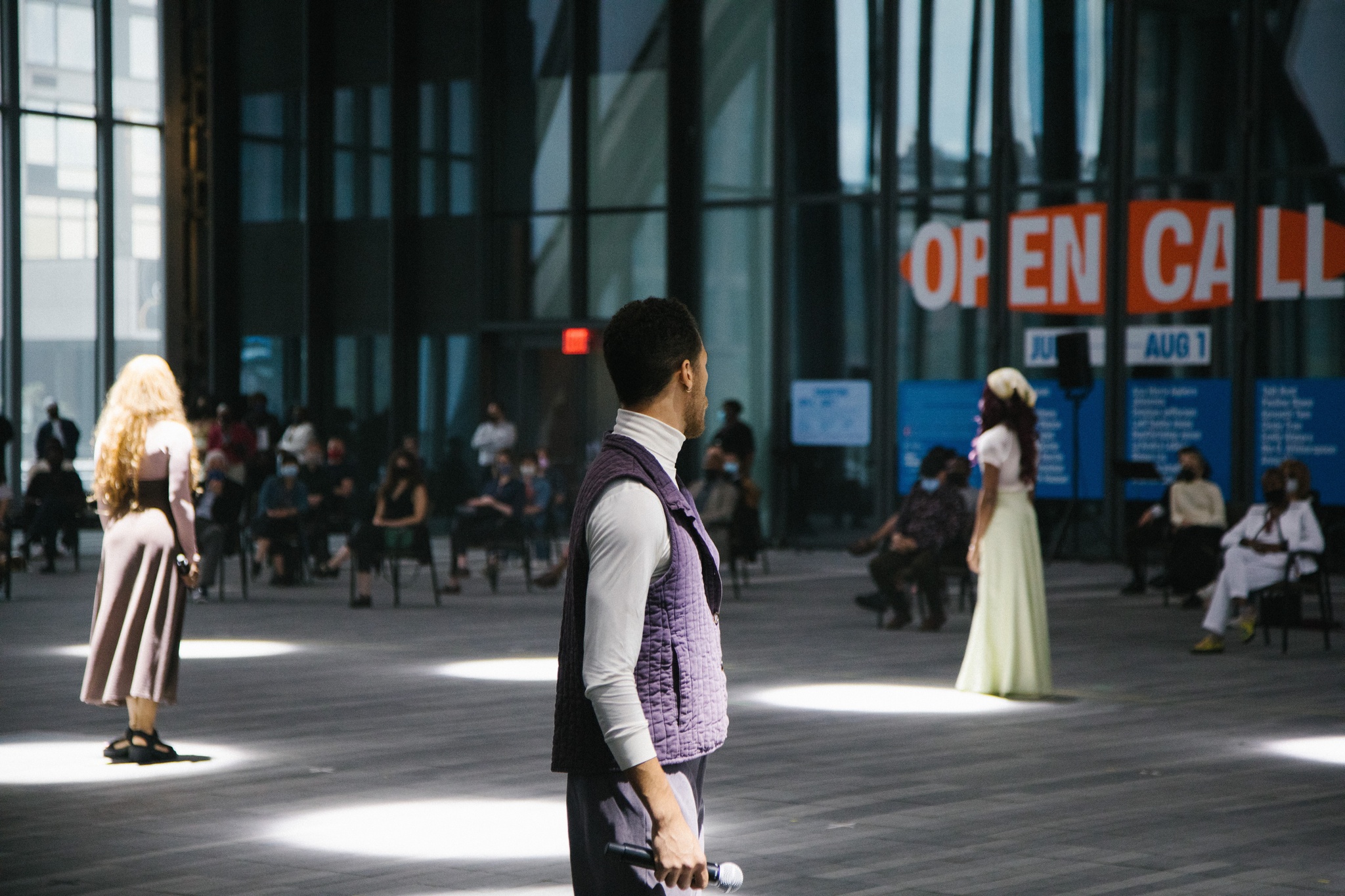 Three performers stand at a distance from one another, each illuminated by a spotlight. In the background, audience members sit in pairs of two.