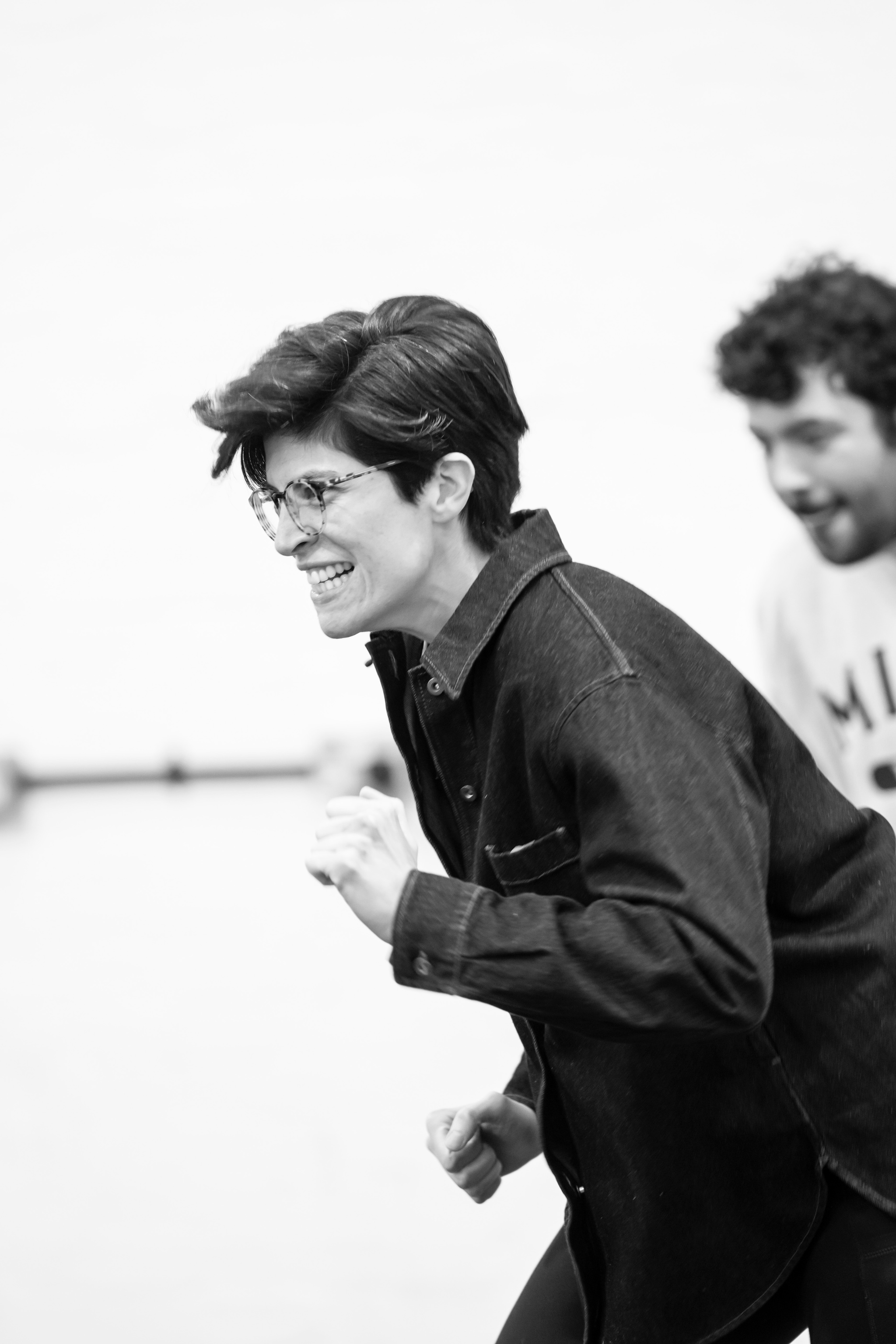 Actor Chloe Fenwick-Brown, a white person with short brown hair parted to the side, is in rehearsal. Chloe bends over in movement with one hand clutched in a fist.