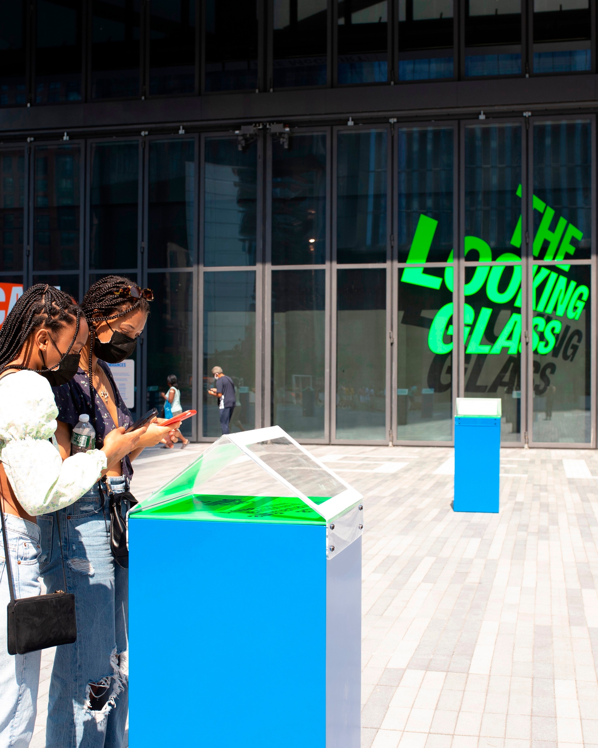 Two people with long braids lean over a bright blue plinth used to activate the exhibition The Looking Glass on a sunny outdoor plaza