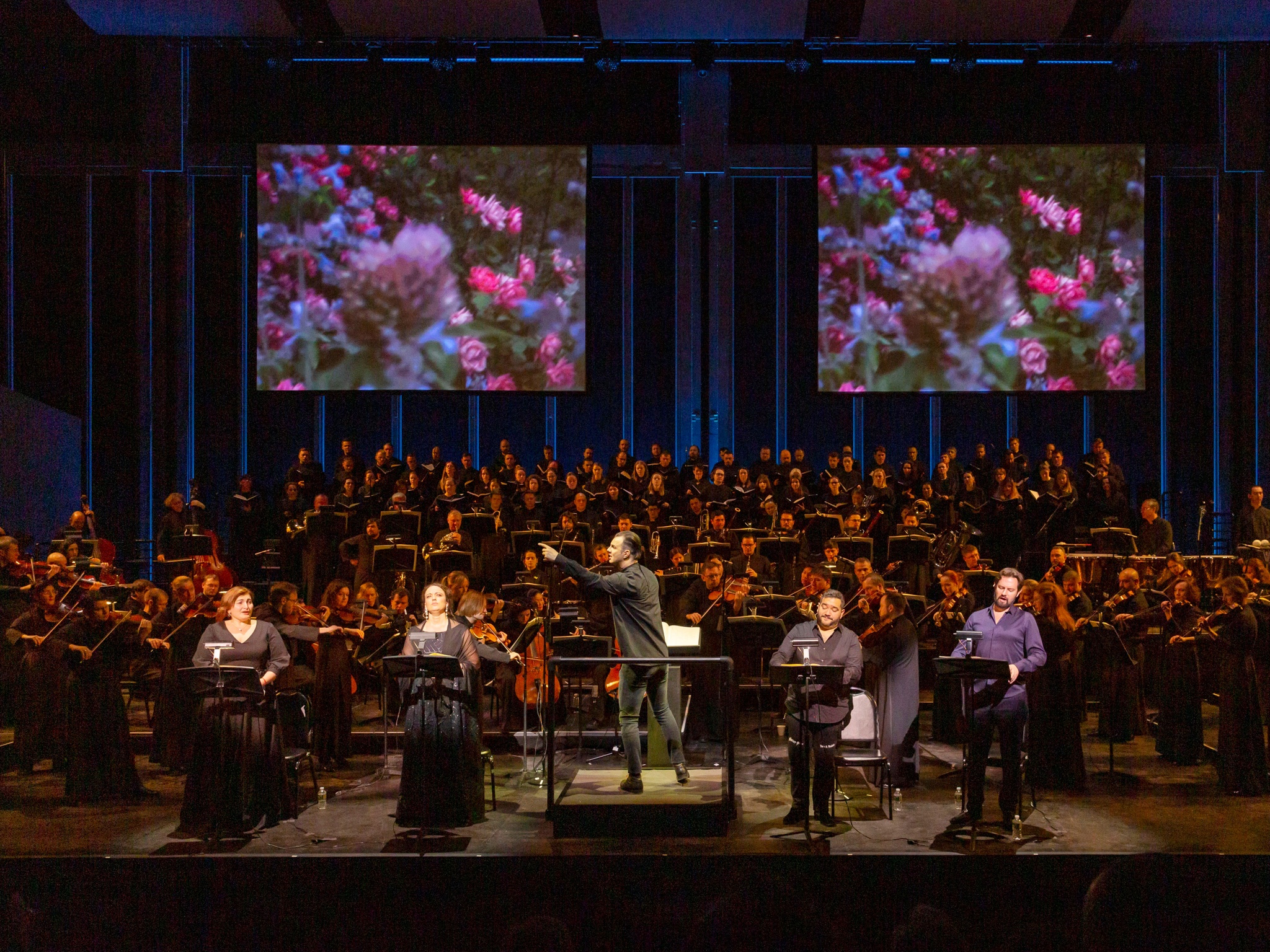 Teodor Currentzis conducting an orchestra in front of two large screens.