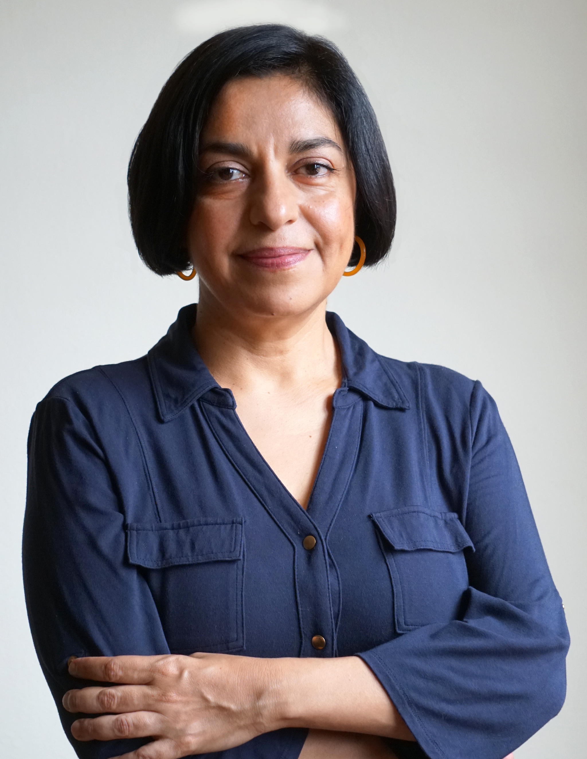 A photo of Claudia Norman with arms crossed across her torso, smiling at the camera. Norman wears a blue button-down blouse and has short, jaw-length dark hair. 