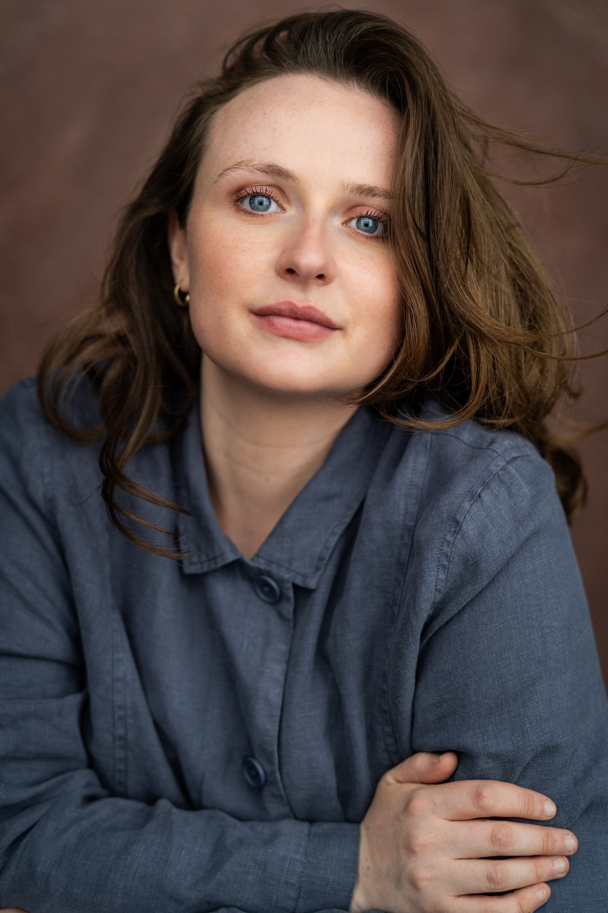 A headshot of actor Ayla Wheatley, a white woman with long brown hair swept to one side, and that falls over her left eye. She leans forward slightly looking directly at us intently. 