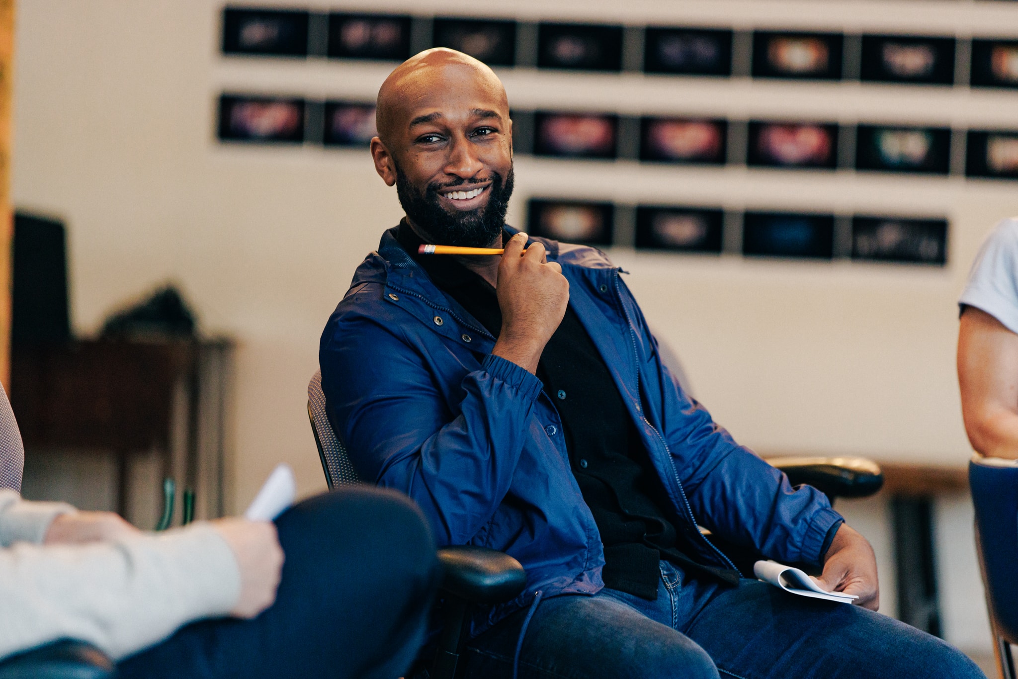 Actor Donald Webber Jr., a black man with a beard and shaved head, sits in a chair during a rehearsal, looking to the side with a wide grin on his face. He wears a royal blue raincoat and holds a pencil up in one hand. 