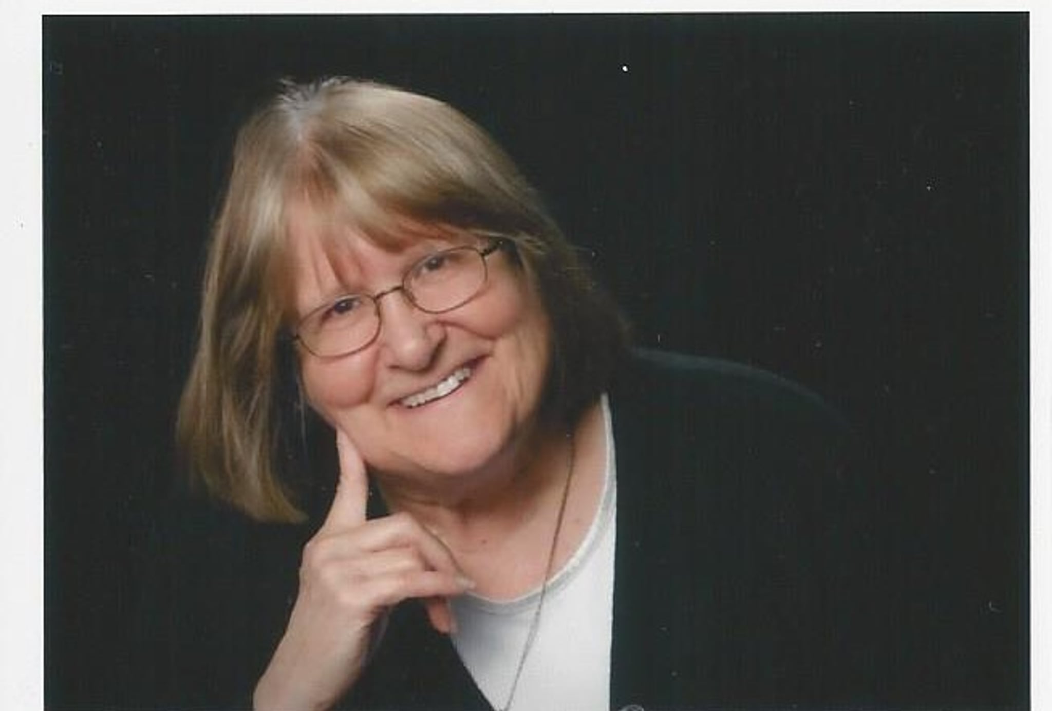 A white woman with blond hair parted down the middle tips her head to the side and holds one hand to her chin. 