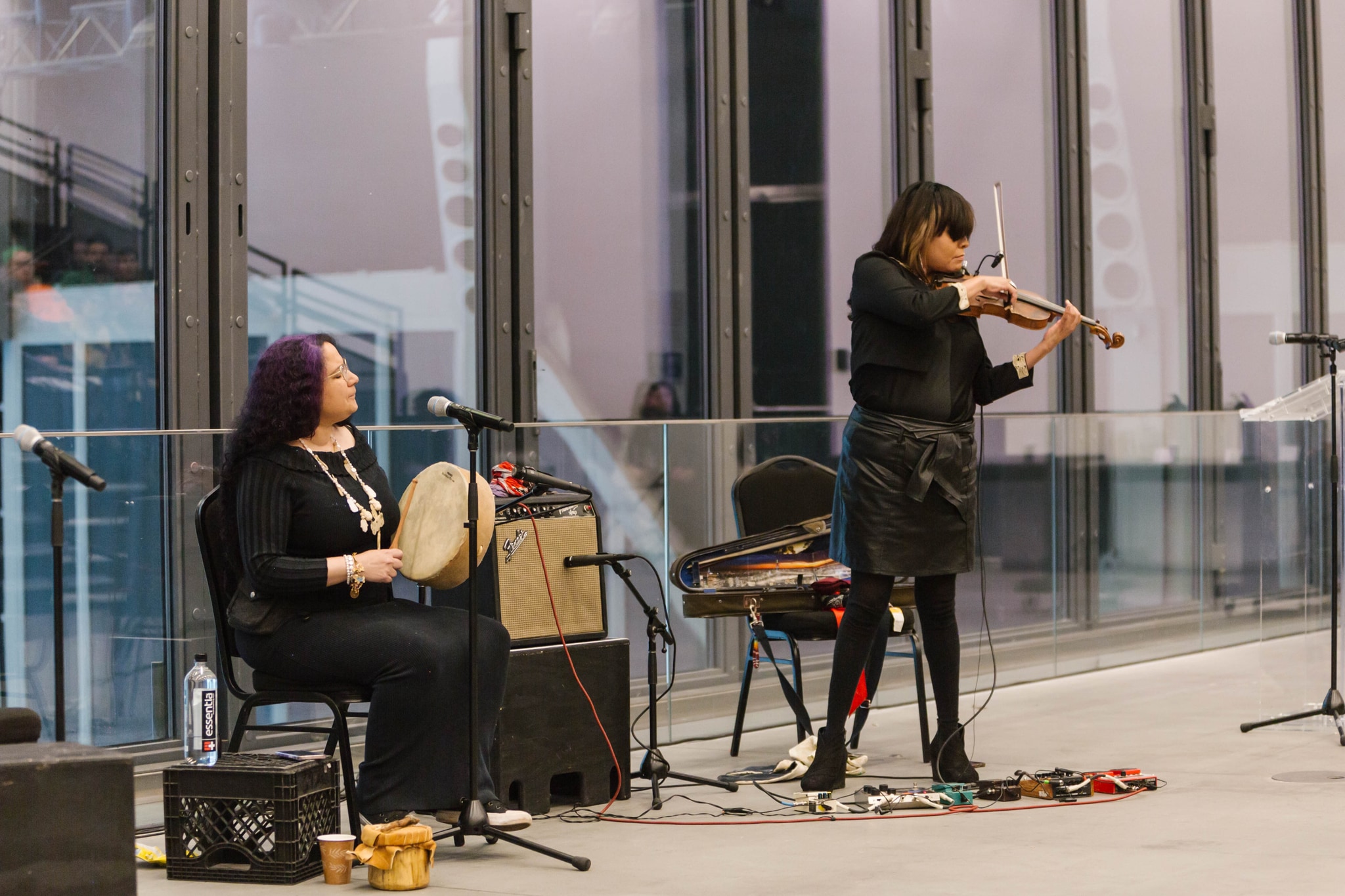 Musician Jennifer Kreisberg sits with a handheld drum on her lap while violinist Laura Ortman stands to her side playing her instrument. Both women wear all black. Jennifer looks to her side to watch Laura play. 