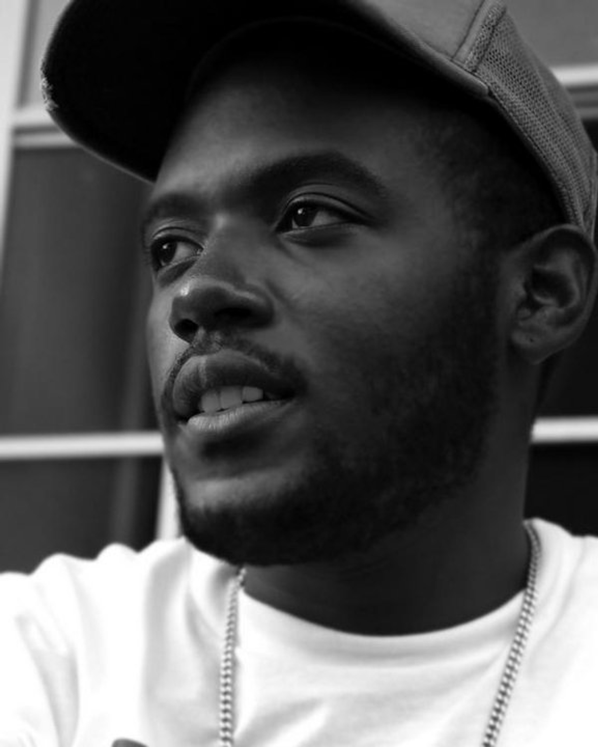 A black-and-white portrait of artist David Sainté in a baseball cap, white shirt, and wearing a chain around his neck