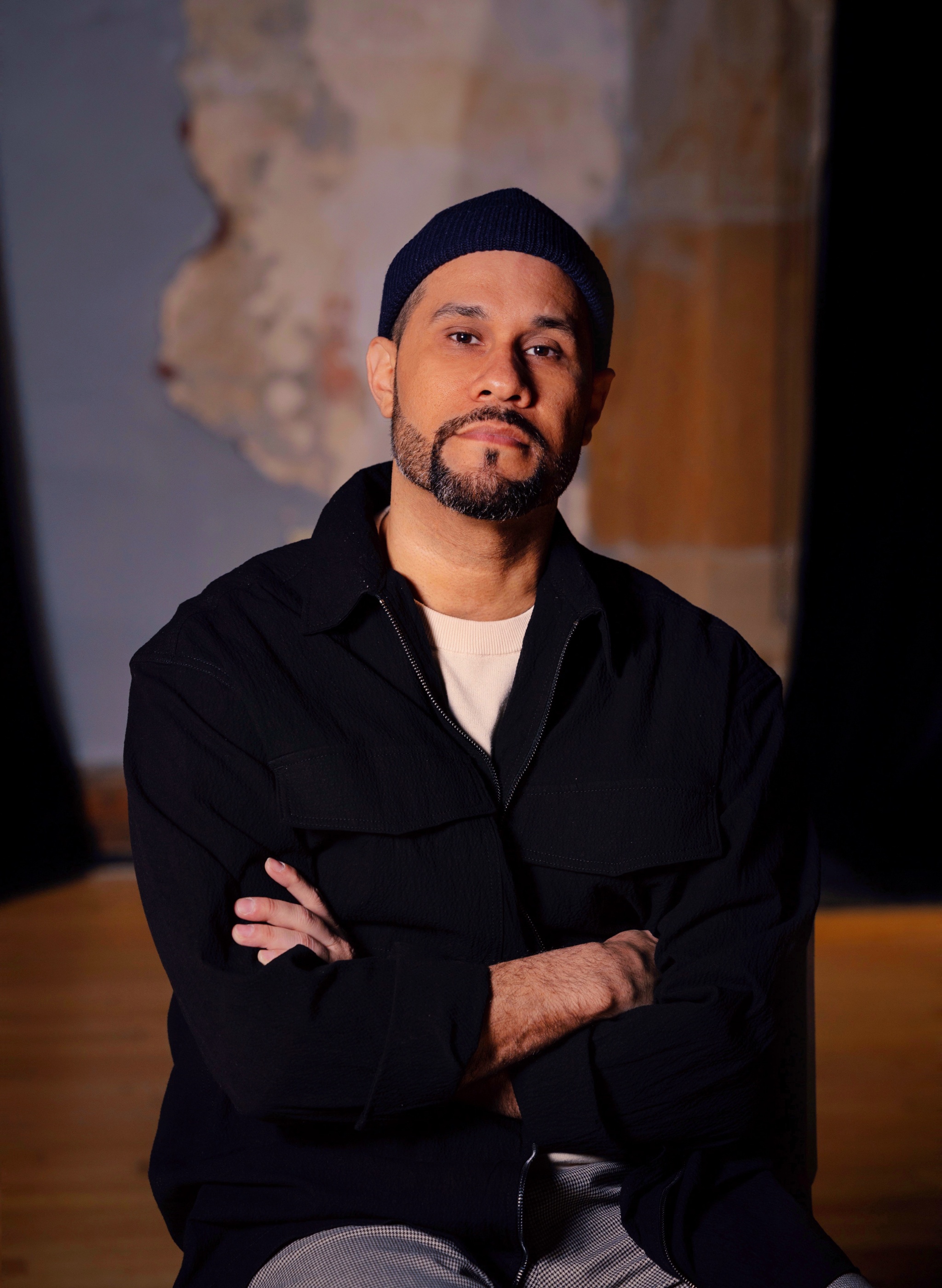 A portrait of artist Rudi Goblen. Rudi is a Nicaraguan man who sits with arms crossed over his chest, facing us and looking directly at us. He has a trimmed mustache and beard and wears a royal blue cap and jacket zipped up over a white t-shirt. 