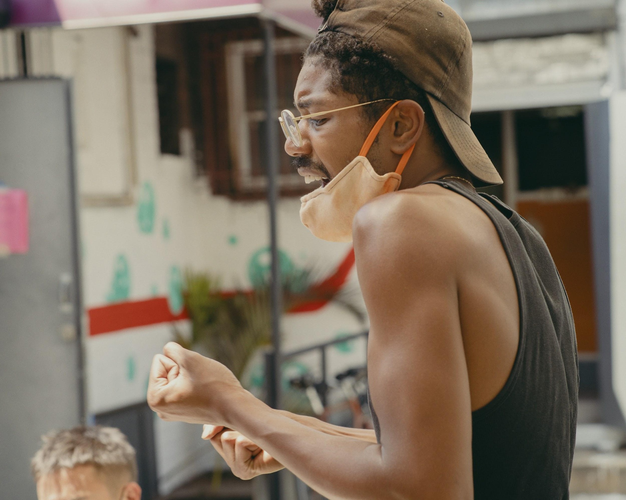 A photo of the artist DonChristian Jones taken from the side. Jones wears a black tank top, a mask around his chin, glasses, and a backwards baseball cap while gesturing with his fists.