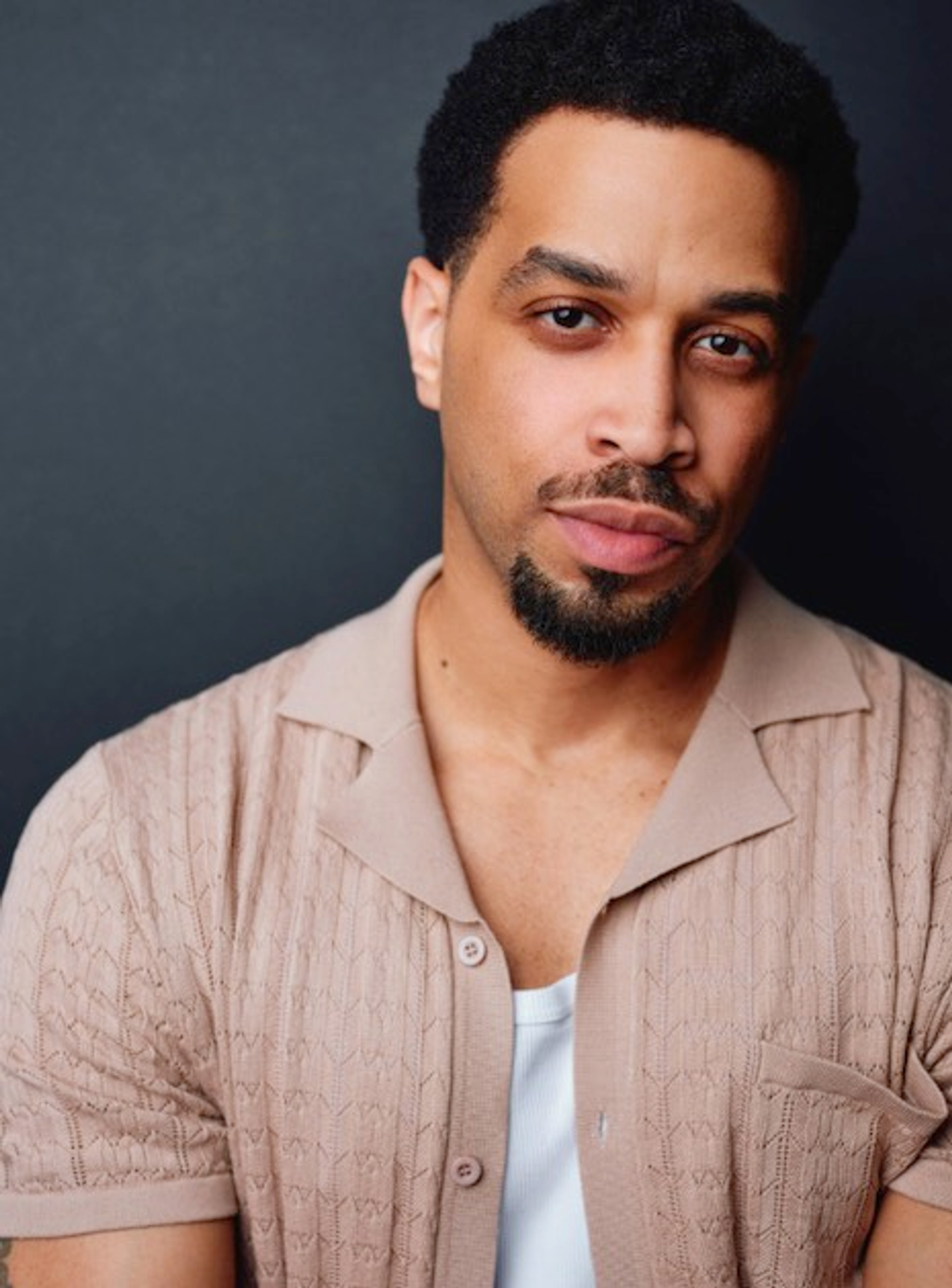 A headshot of actor Stephen Cofield, Jr. Stephen is a Black man with light brown skin who poses against a dark gray backdrop. He has a mustache and goatee beard, wears a beige short sleeve button down shirt open over a white tank top, and looks intently at us. 