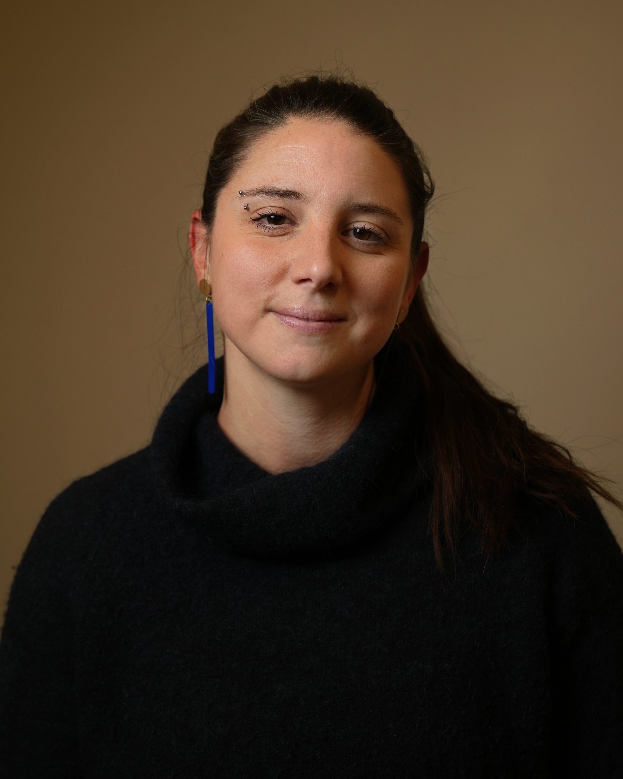 A portrait of Frances Caperchi against a neutral brown background. Frances has long dark brown hair swept behind her head. She looks directly at us with a slight smile on her face.