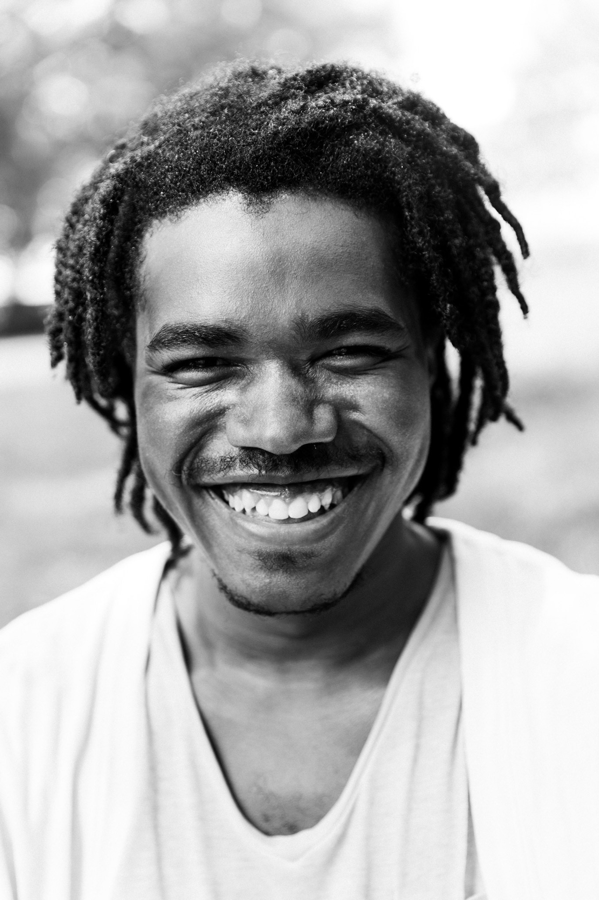 A portrait of a smiling Black man wearing a white v-neck t-shirt. He has a mustache across his upper lip and a beard that lines the area just under his chin. 