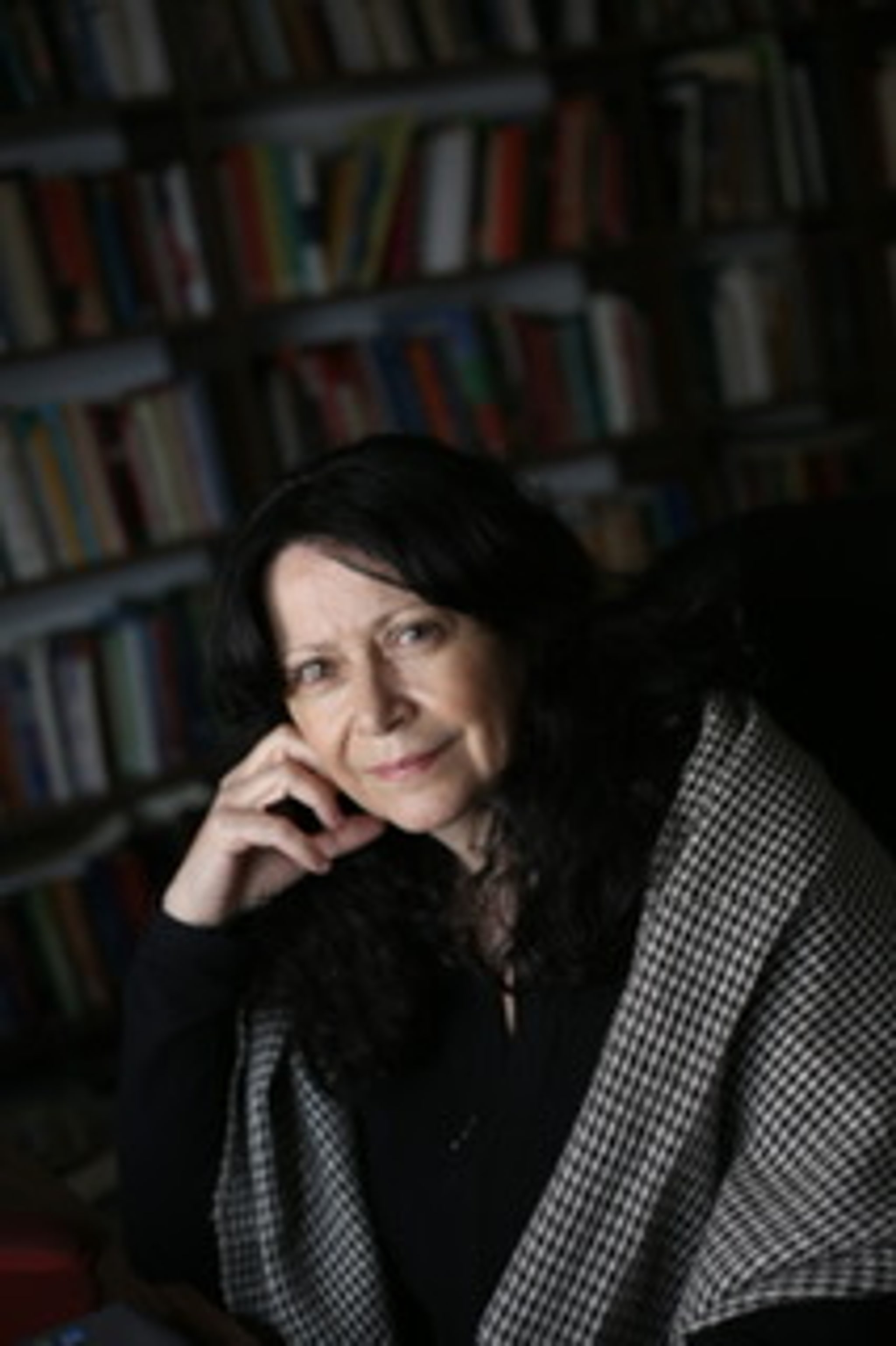 A woman with dark brown hair sits with her chin propped on one hand. She wears a checkered wrap around her shoulders.