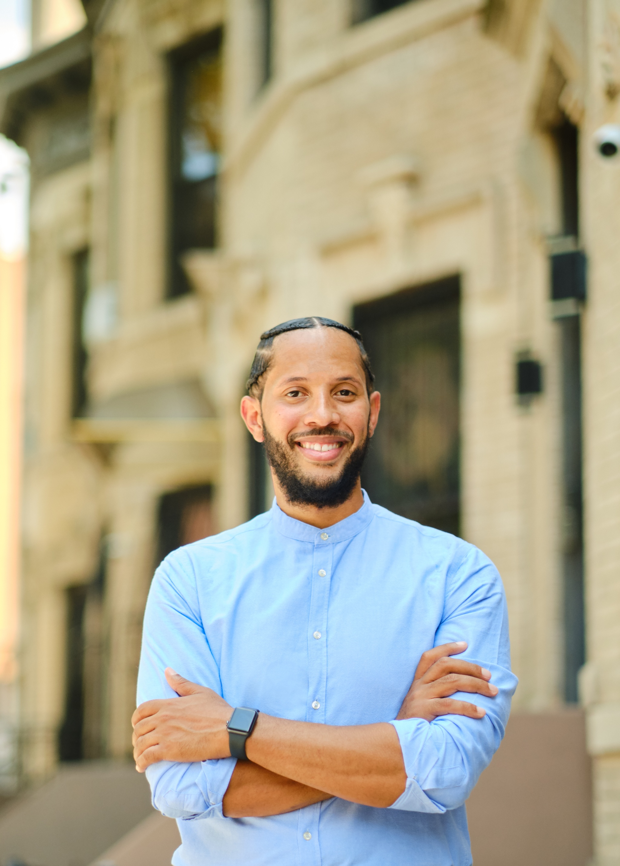 André Richardson poses with arms crossed in front of a row of building façades blurry in the background. He has a beard and wears a blue button-down shirt. 