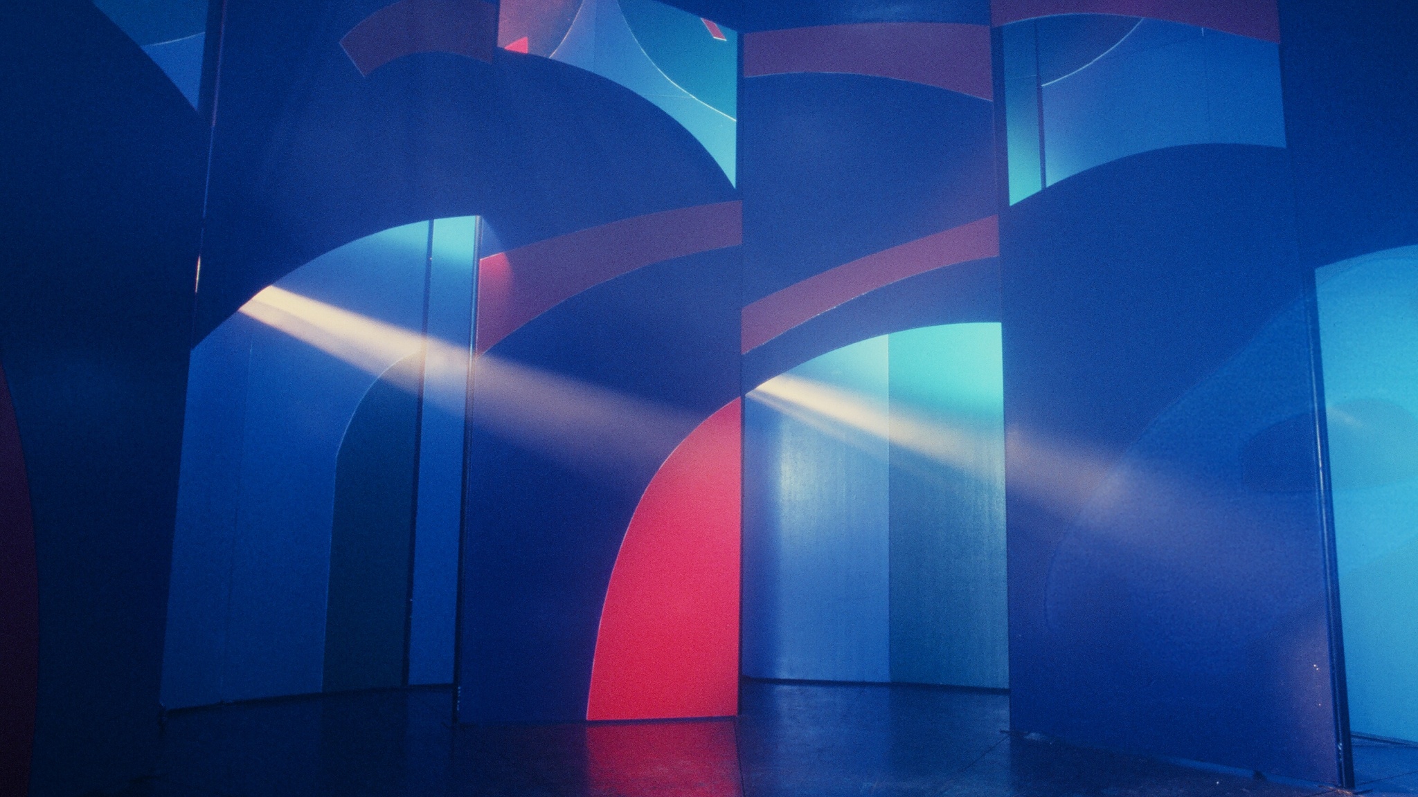 The interior of a carnival attraction by artist David Hockney. The space is moodily lit in dark blue and pink lights. A ray of bright white light shoots across the room from high above. 