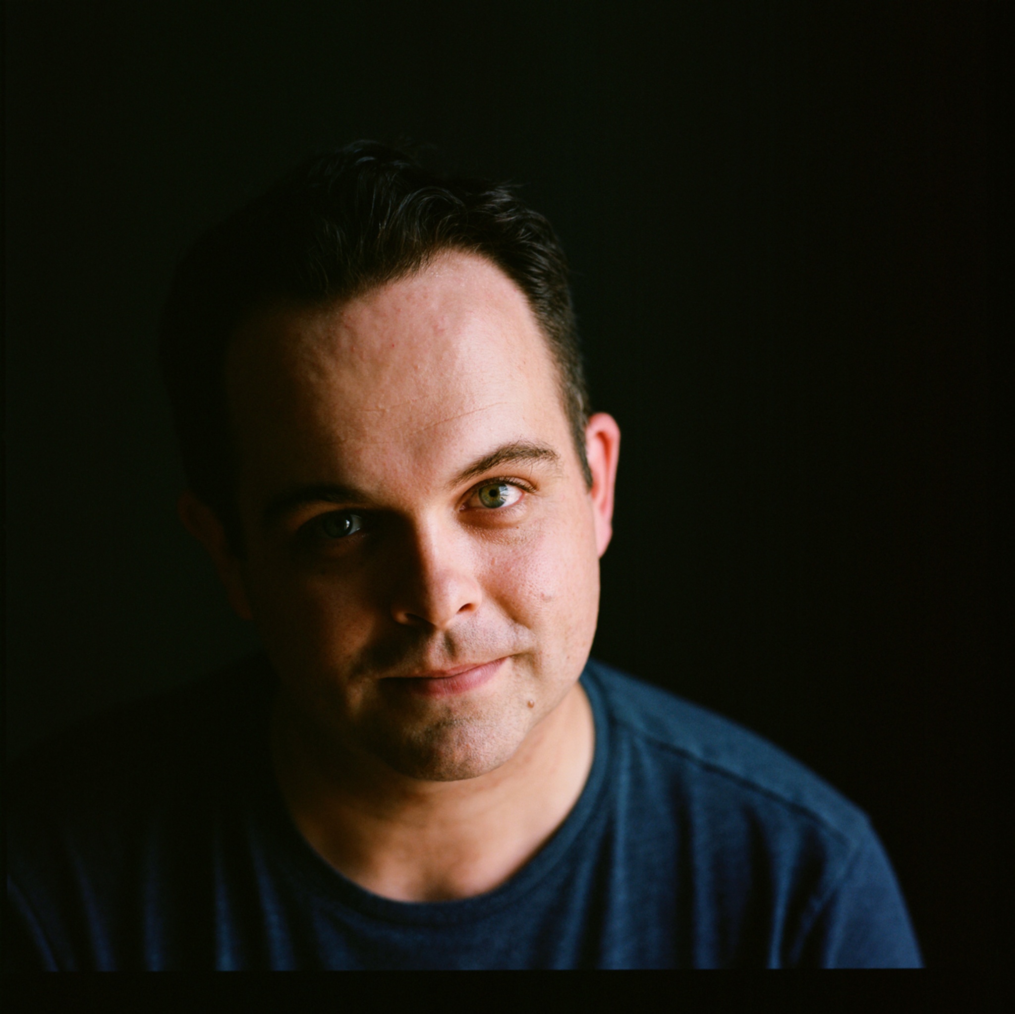 A photo of Andrew Kircher against a dark background. Kircher wears a dark blue shirt and the right side of his face is in shadow. 