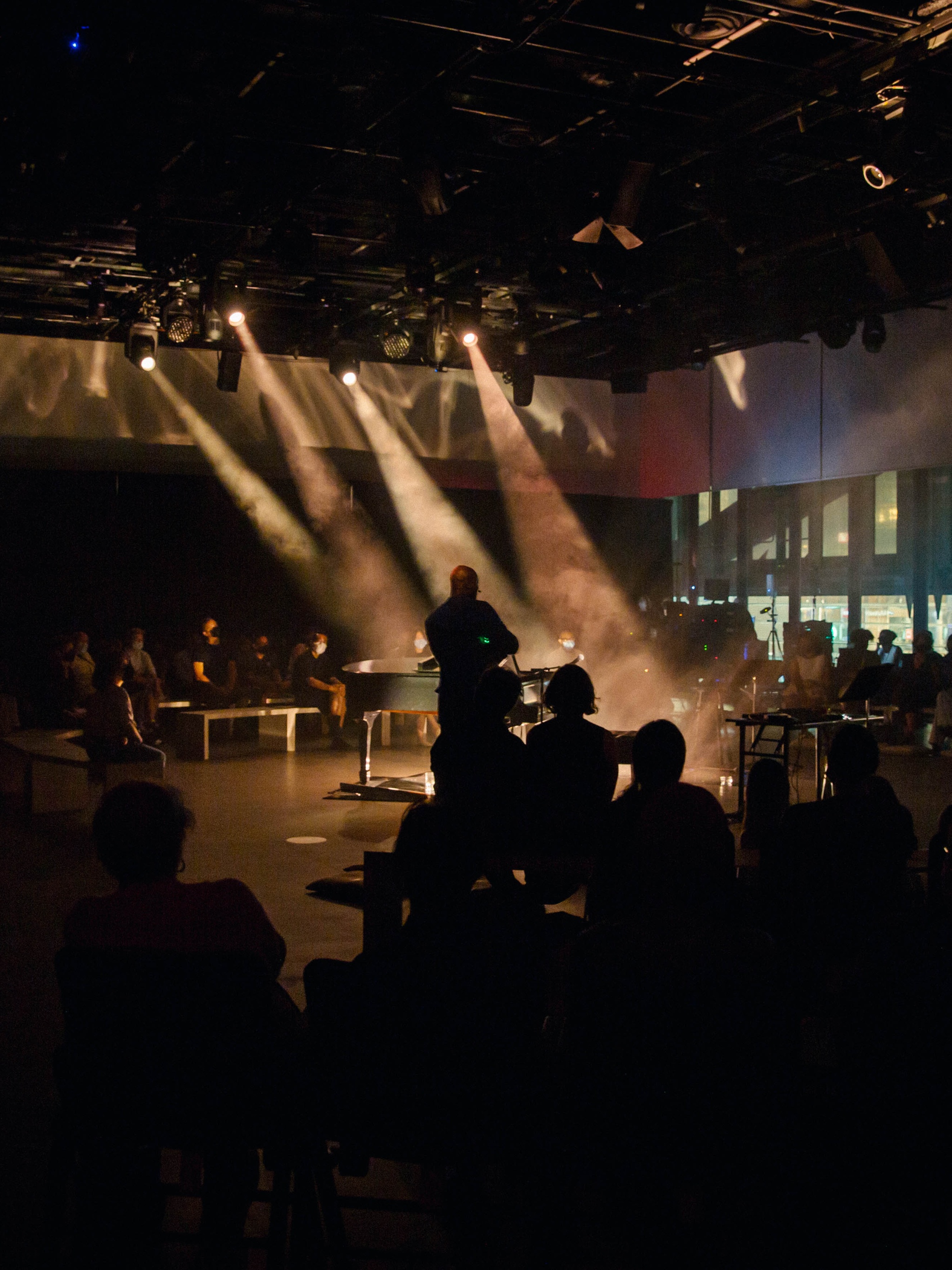 The artist Justin Hicks whose face and features are obscured stands at the center of an audience surrounding him. The space is stands in is lit dramatically by stage lights that send beams of light through the foggy air.