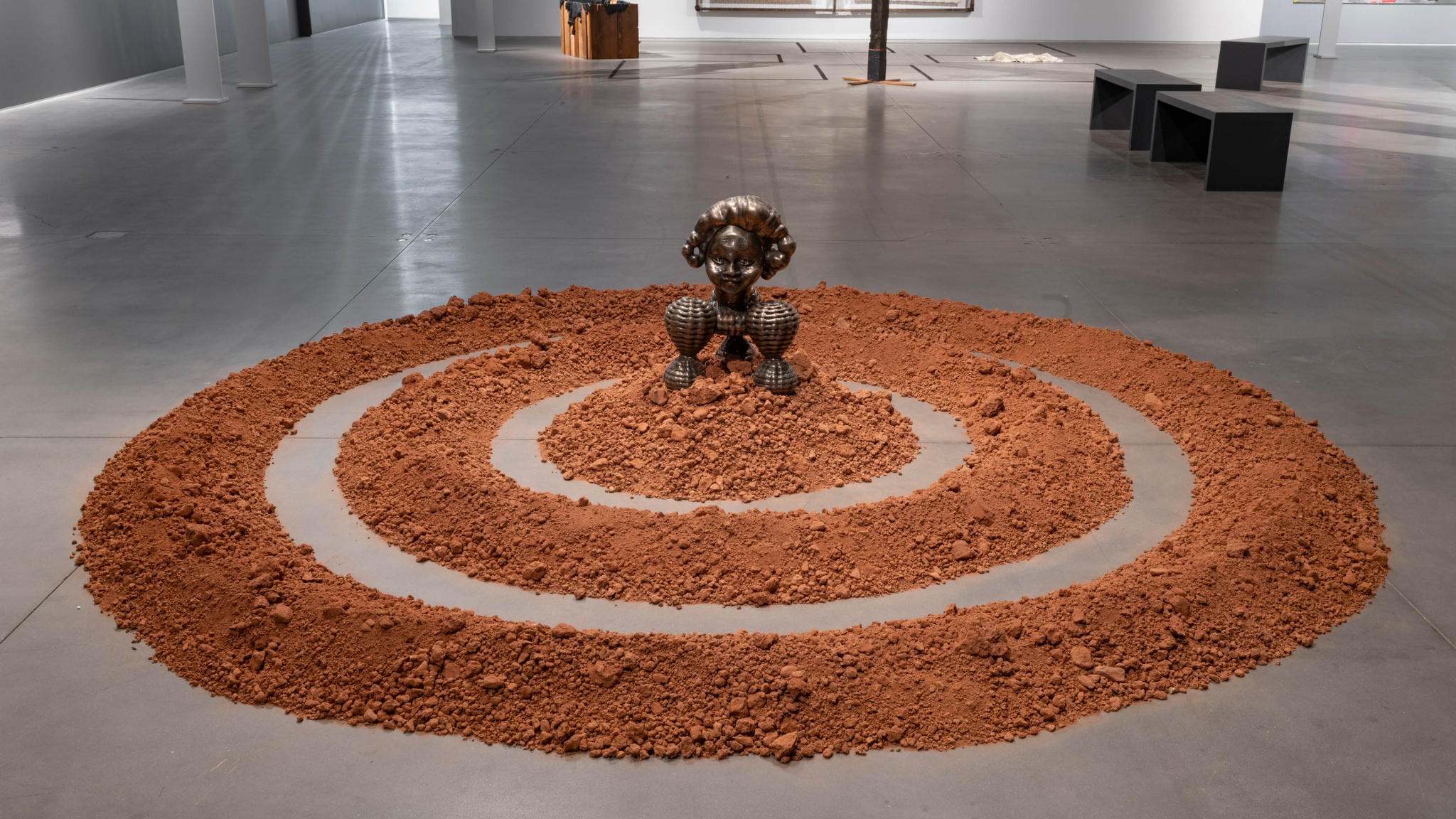 A wide sculptural artwork on a gallery floor. It consists of two concentric rings of red dirt. At the center on top of a mound of the dirt sits a bronze sculpture inspired by the Benin Bronzes, historical artworks from Benin, present-day Nigeria.