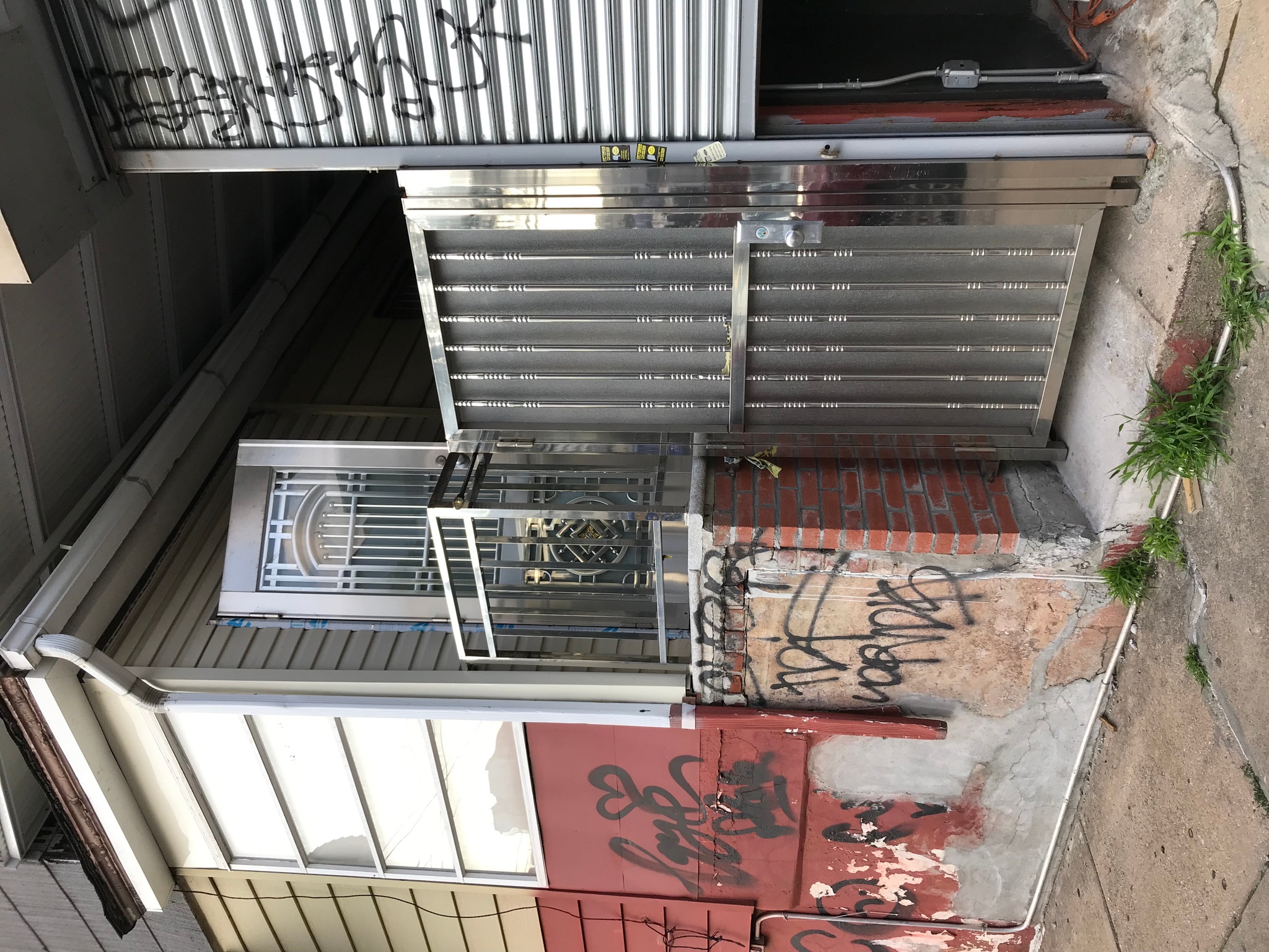 A corner of a house with a stainless steel gate closed upon the sidewalk alley beside a porch elevated a half story above street level. The porch has a stainless steel front door that is half open to the house, though it blocks any view inside the structure.