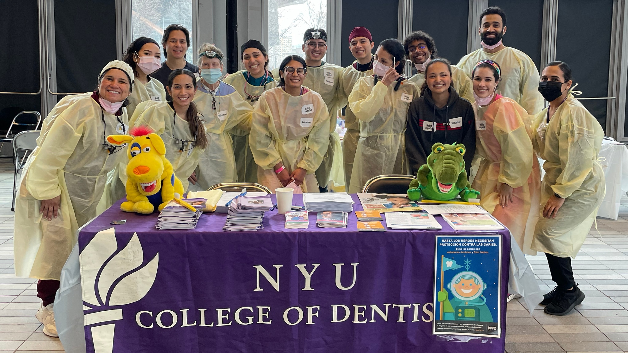 A group of 15 dentists in yellow protective gear gather for a group picture behind a table draped in a purple cloth emblazoned with "NYU College of Dentistry"