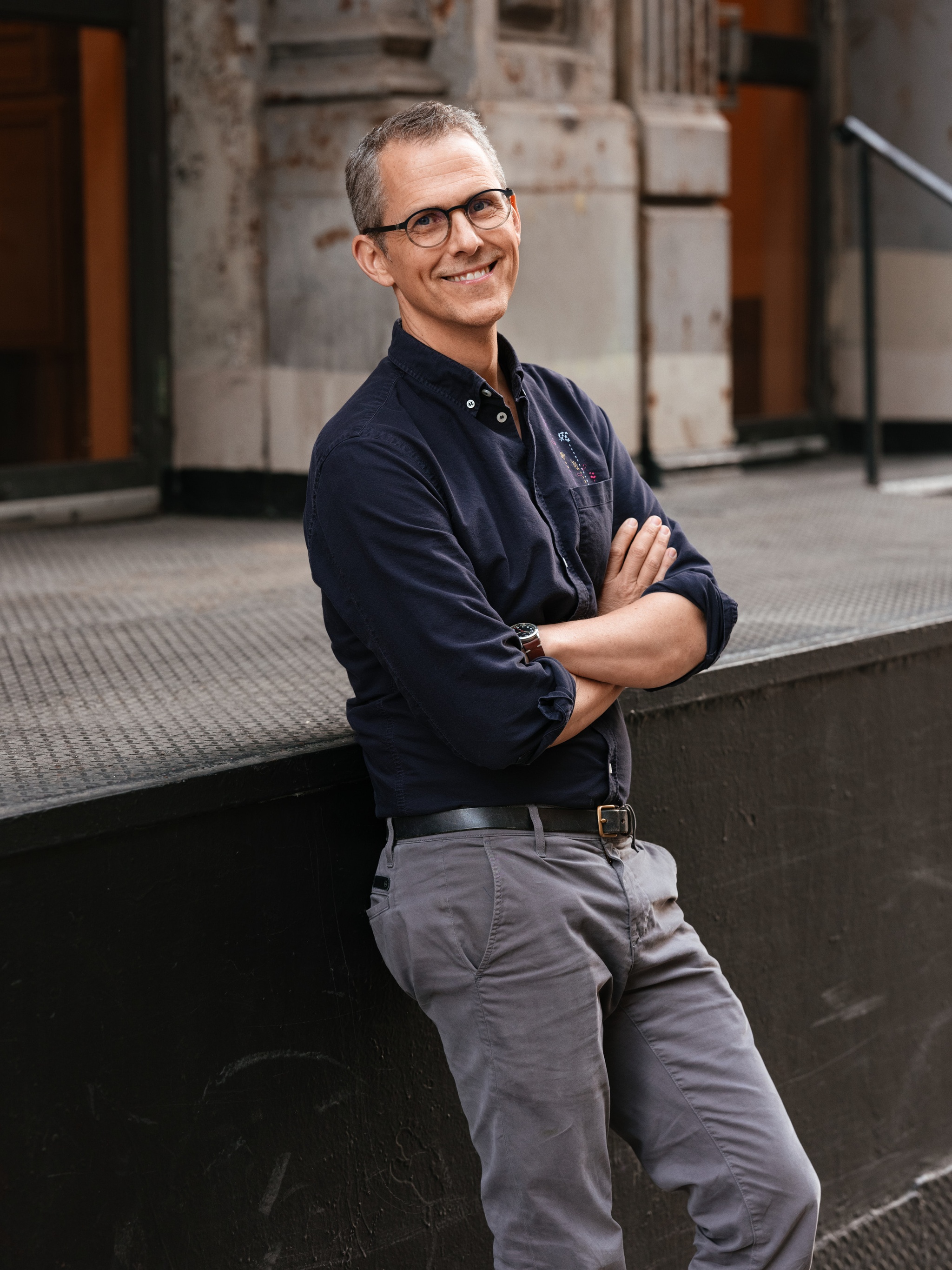 A white man in a blue button down shirt tucked into gray jeans leans against a metal platform on a city sidewalk.