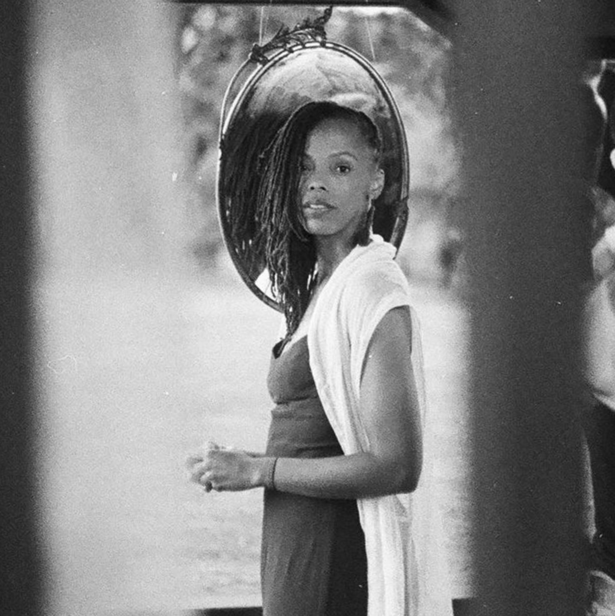 Jillian Walker, a Black woman, looks at us in a black and white portrait. She is seen from the side turning her head to look at us. Her locs fall over the right side of her face and she wears a long white duster. 