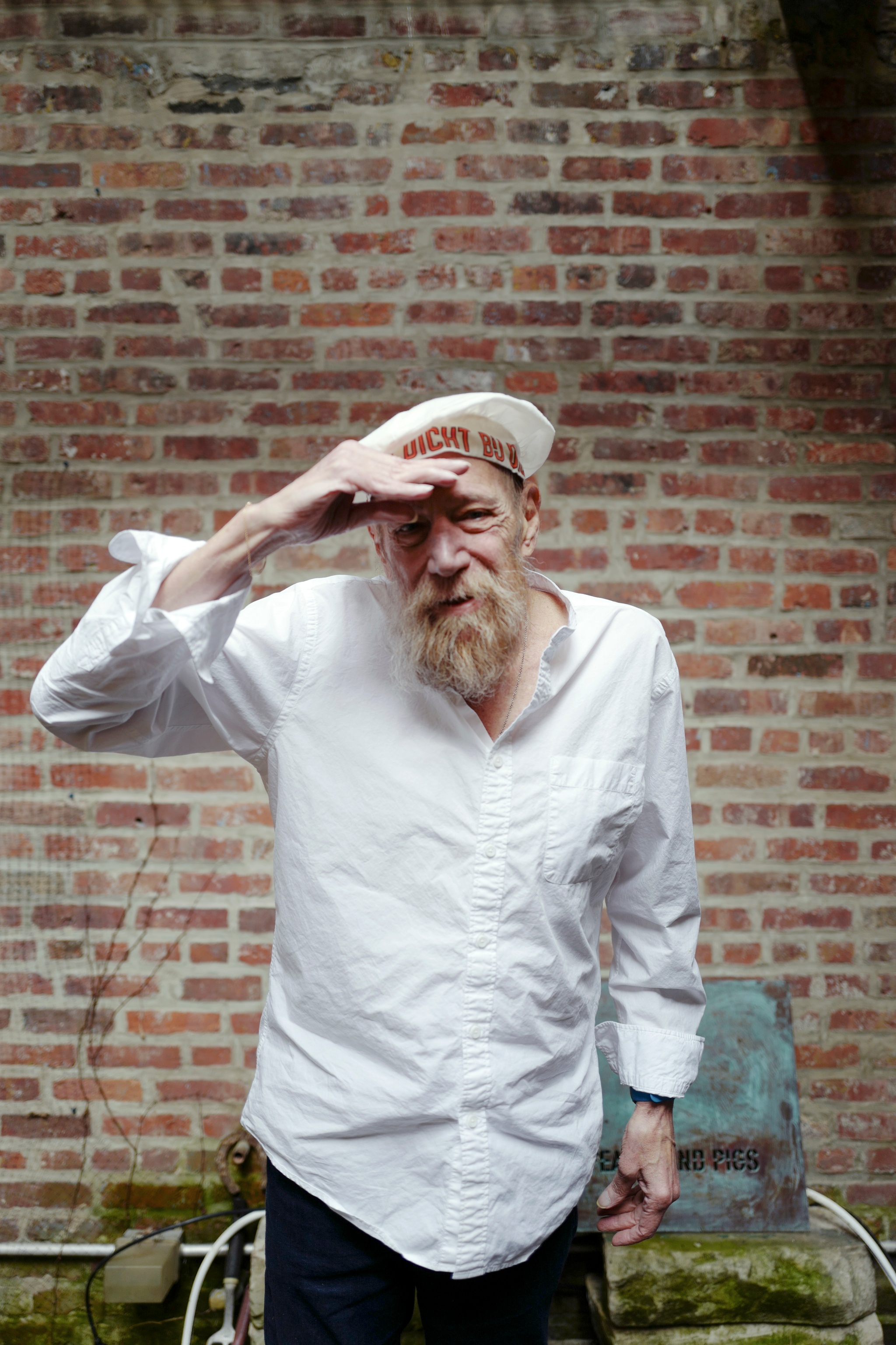 A portrait of artist Lawrence Weiner, an older white man who wears a rumpled white button down shirt and a white cap. He has a mid-length red-grayish beard and looks directly at us with smiling eyes, one hand held up above his brow as if shielding his eyes from the light.