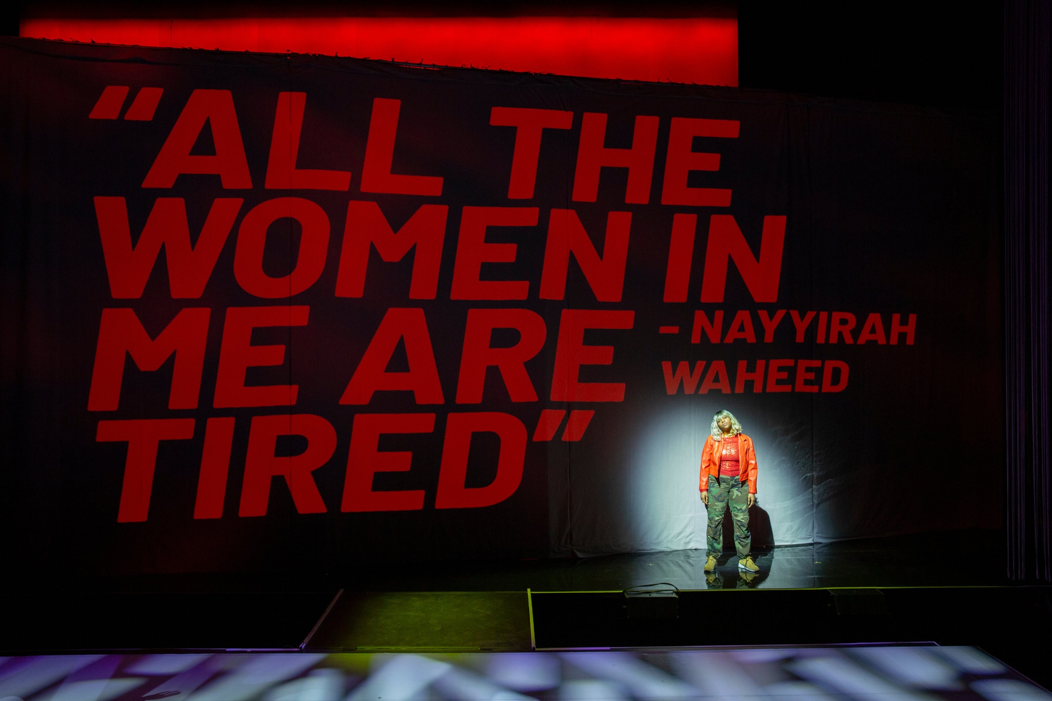 LATASHÁ, a black woman with bleached blond hair to her shoulders, stands in a spotlight on stage. She wears a bright orange jacket and on a curtain behind her are projected the words "All the women in me are tired" in bright red letters.