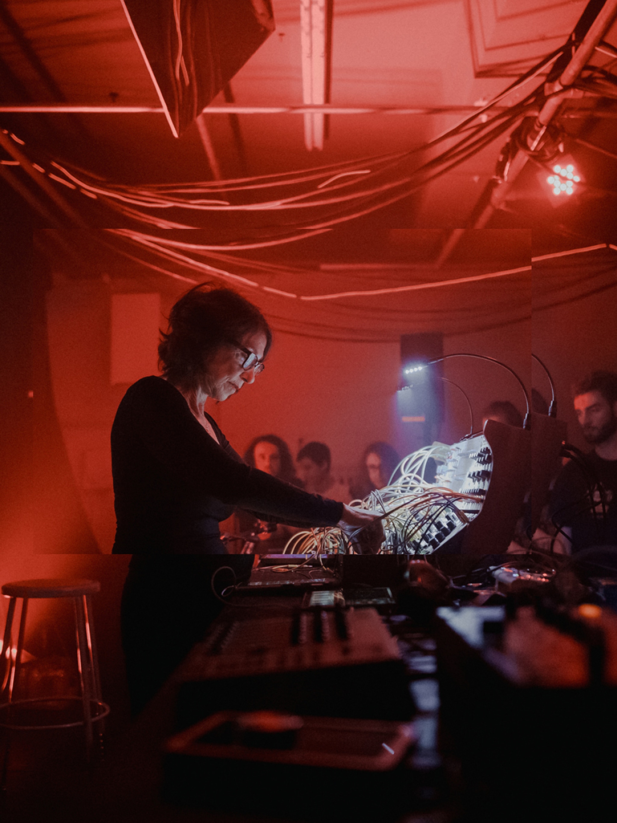 Musician Suzanne Ciani, a white woman with short wavy brown hair, is seen in profile manipulating a sound board. The small space is lit by red light and an audience is gathered on the other side of the sound board. 