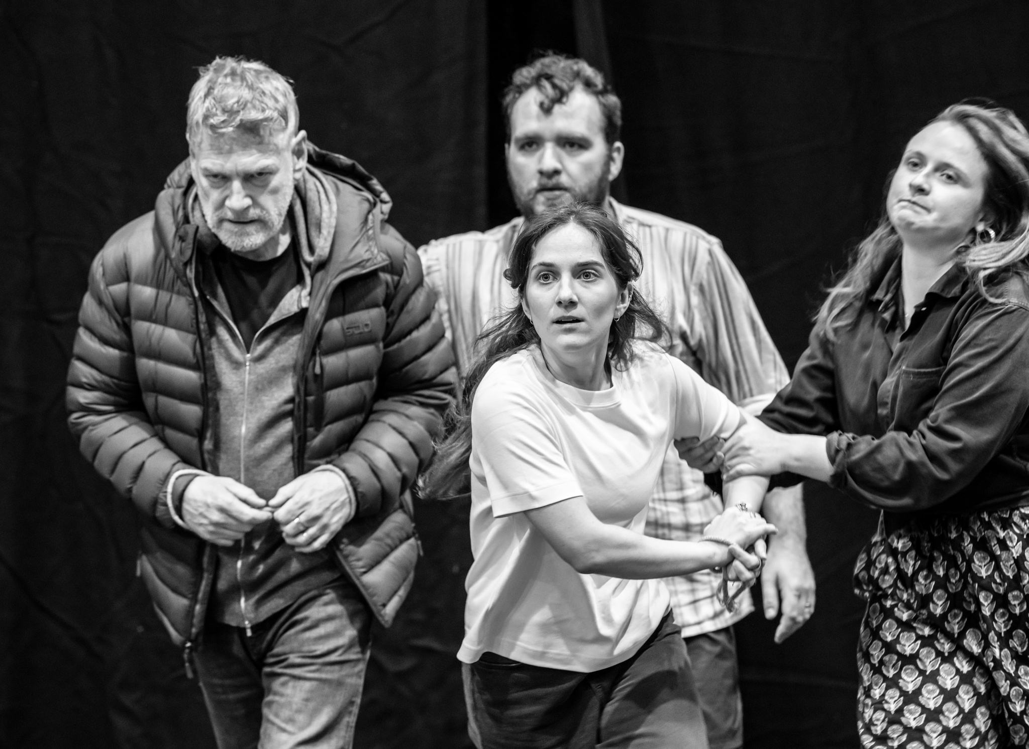 Actors Kenneth Branagh, Hughie O'Donnell, Jessica Revell, and Ayla Wheatley in rehearsal. They stand close together moving forward, with intense looks on their faces. Ayla reaches out to grab Jessica's shoulder, trying to hold her back. 