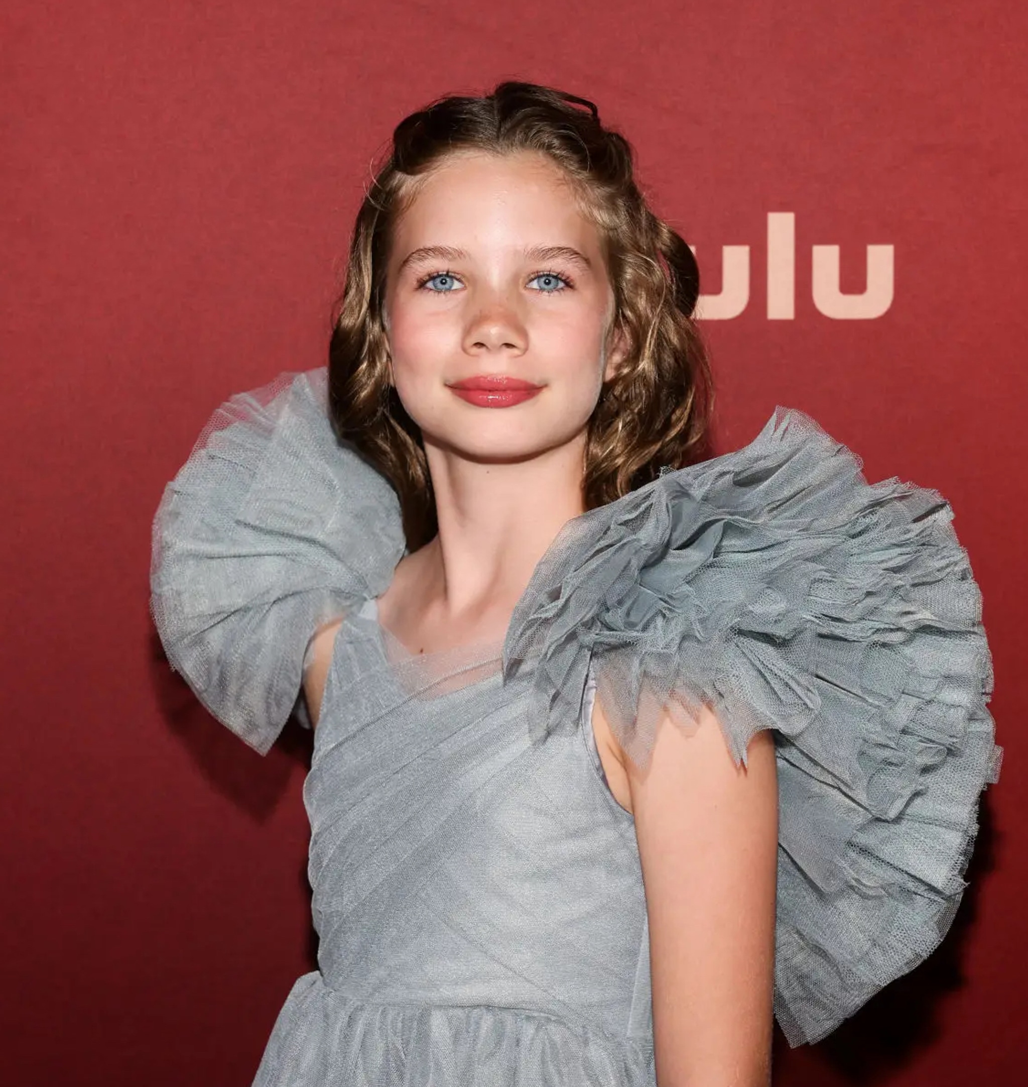 A portrait of Alona Jane Robbins, a young Deaf actor. Alona is a white girl with dirty blond hair. She wears a silver colored gown with puffy shoulders and smiles directly at us, standing against a red step and repeat backdrop.
