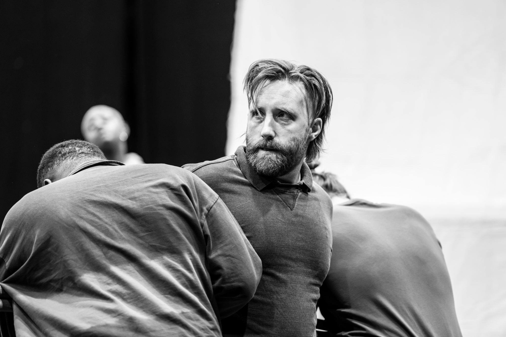 In rehearsal, actor Joseph Kloska, a white man, is restrained by two other actors who hold him from either side. Kloska looks intently to the side, while the other two actors' heads are down and backs turned toward us. 